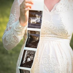 SKY Lens Radosław Nikonowicz - Kobieta w koronkowej sukience ciążowej trzymająca wydruk USG, ukazujący wizerunek dziecka. Delikatne światło, naturalne tło. Sesja w plenerze.