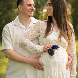 SKY Lens Radosław Nikonowicz - Uśmiechnięta para w plenerze: przyszła mama w koronkowej sukience trzyma buciki niemowlęce, tata obejmuje brzuszek. Romantyczna sesja ciążowa w naturalnym świetle.