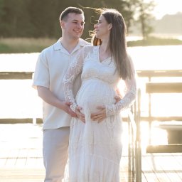 SKY Lens Radosław Nikonowicz - Sesja ciążowa na pomoście: przyszła mama w koronkowej sukni, tata obejmuje brzuszek, oboje patrzą na siebie z uśmiechem, w tle woda i zieleń.