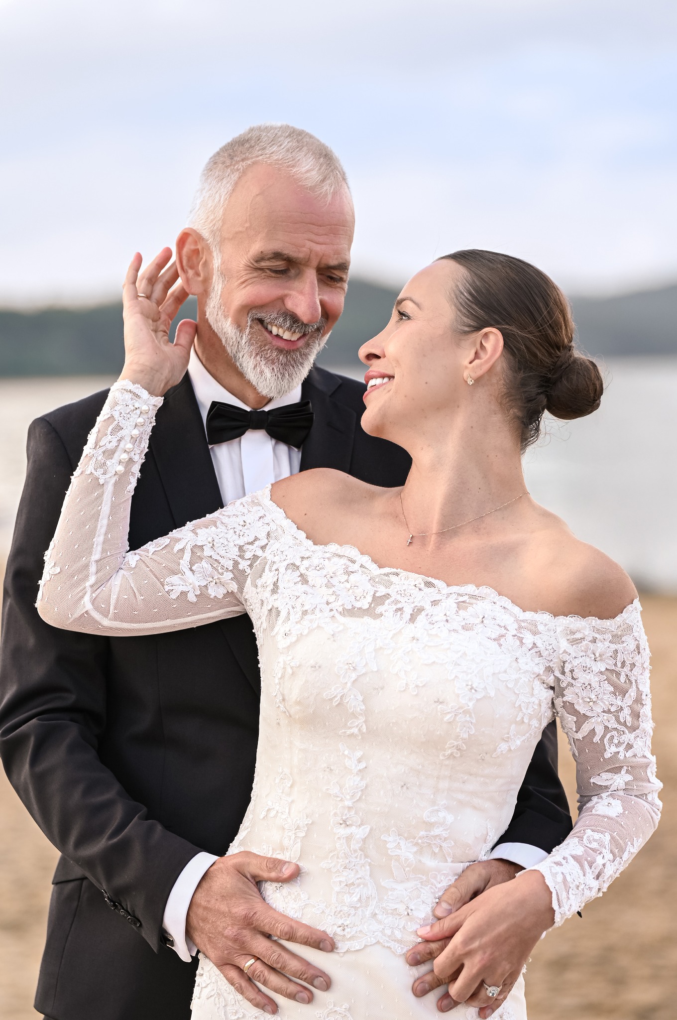 Uśmiechnięta para młoda w objęciach na tle jeziora. Ona w koronkowej sukni z odkrytymi ramionami, on w czarnym smokingu z muszką. Delikatne światło podkreśla ich radość.