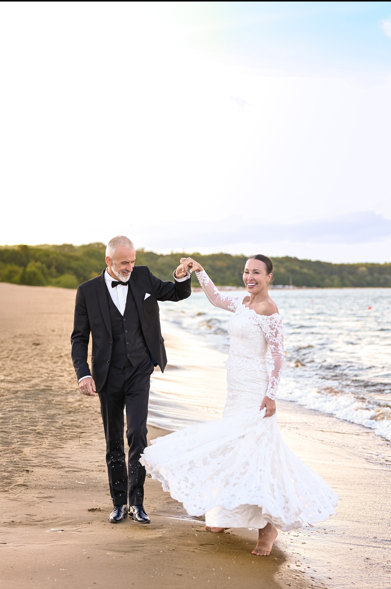 Para młoda, on w smokingu, ona w koronkowej sukni, tańczą boso na plaży o zachodzie słońca. Uśmiechnięci, trzymają się za ręce, w tle woda i zieleń.