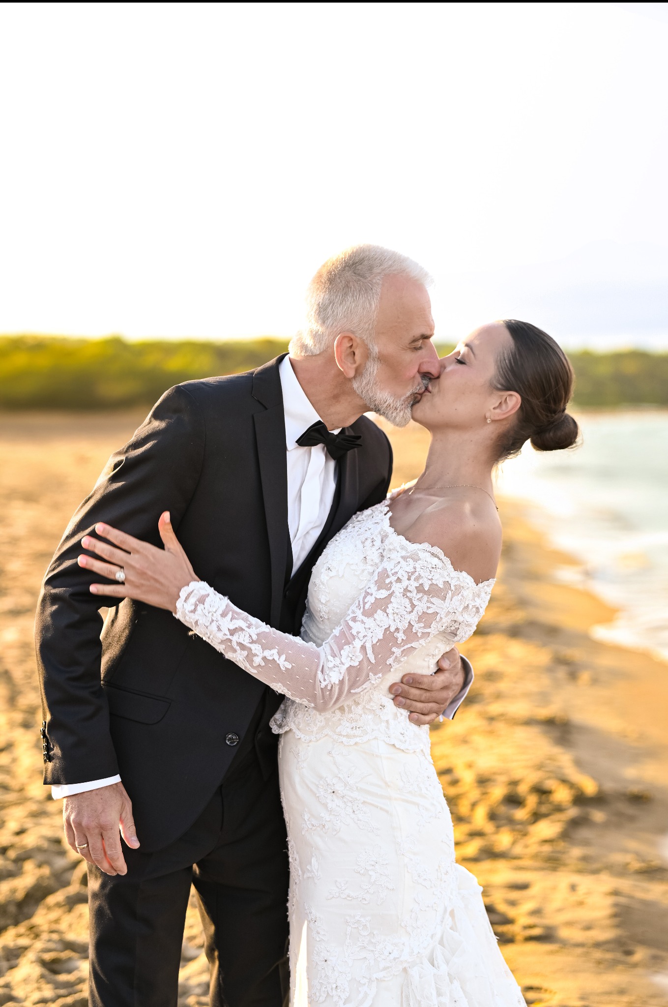 Para młoda w objęciach, pocałunek na plaży o zachodzie słońca. Elegancki pan młody w smokingu, panna młoda w koronkowej sukni z długim rękawem. Romantyczna atmosfera.