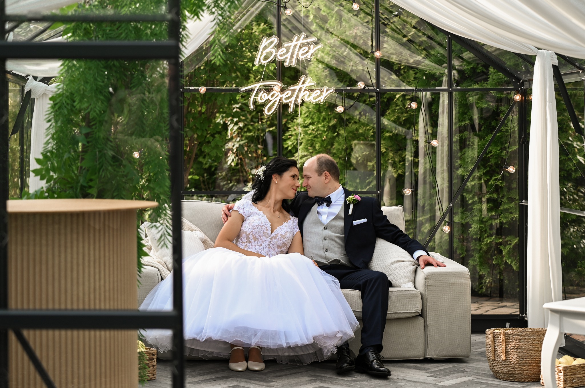 Para młoda siedzi na sofie w szklarni z napisem 'Better Together' z żarówek. Romantyczna sesja ślubna w plenerze, dekoracje ślubne, naturalne światło.