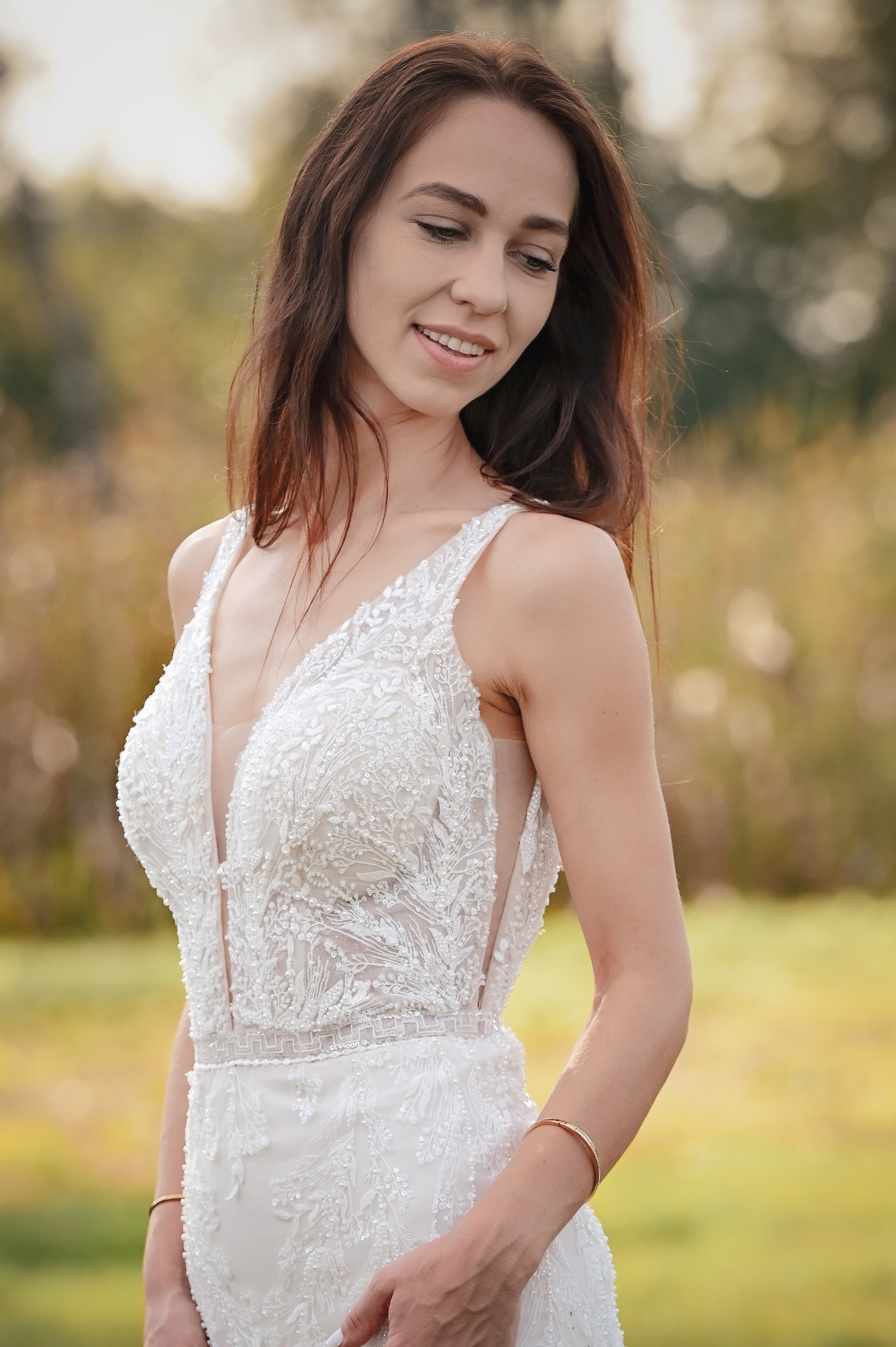 Uśmiechnięta panna młoda w koronkowej sukni z głębokim dekoltem, pozująca na tle rozmytej zieleni i złota jesiennego krajobrazu. Delikatny makijaż i złota bransoletka.