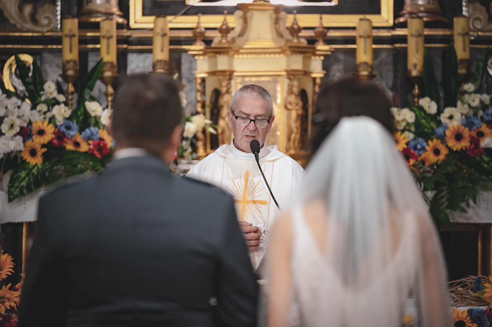 Uroczystość ślubna w kościele: ksiądz w ornacie wygłasza kazanie, w tle para młoda i dekoracje kwiatowe. Delikatne światło podkreśla sakralny charakter.