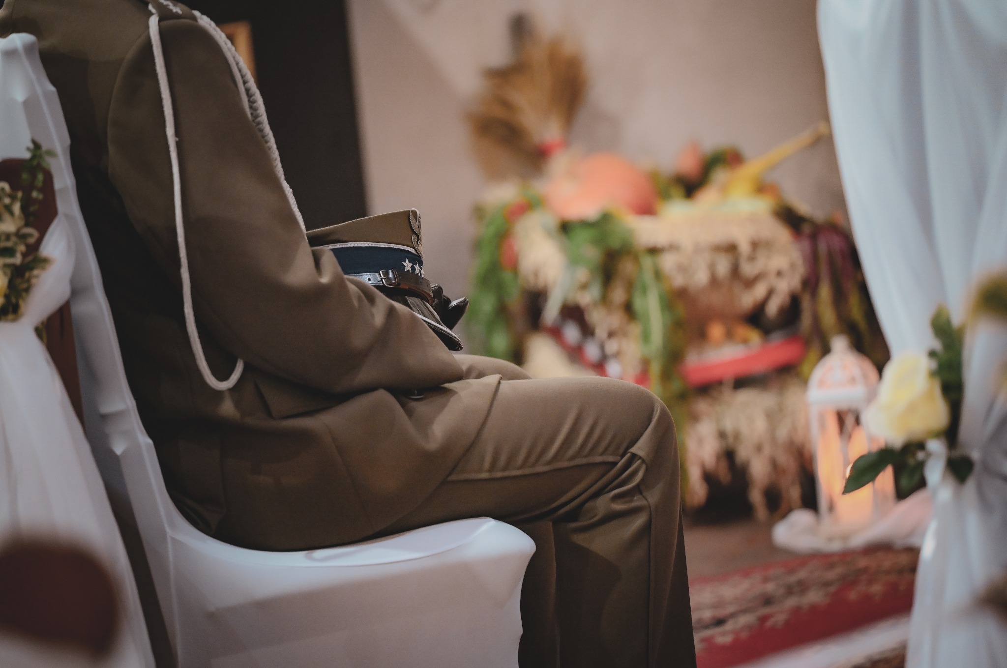 Siedzący mężczyzna w mundurze na weselu, z czapką na kolanach. Dekoracje kwiatowe i lampiony w tle, miękkie światło.