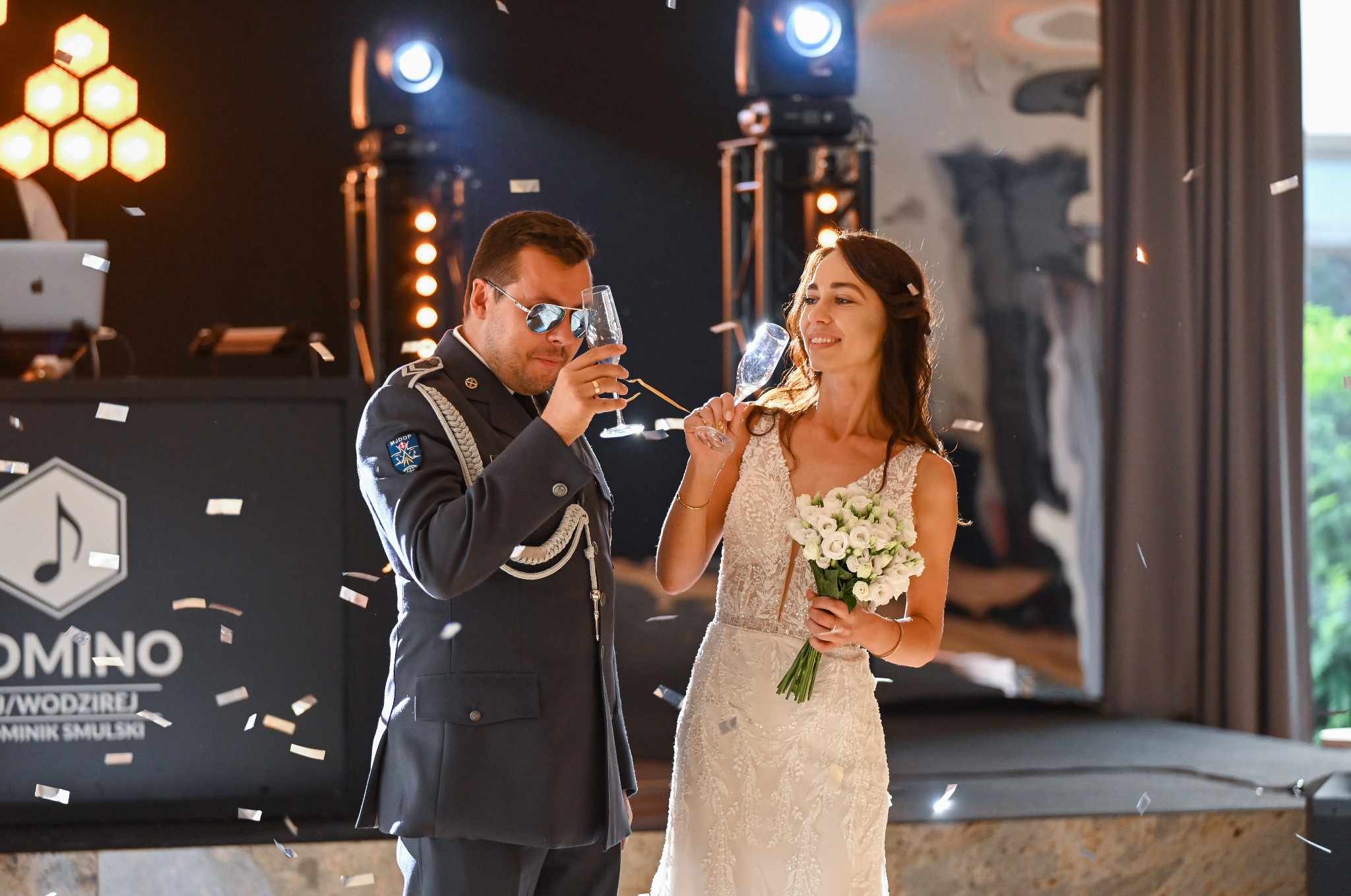 Para młoda wznosi toast szampanem na tle sceny z konfetti. Pan młody w mundurze, panna młoda z bukietem. Uśmiechnięci, w radosnej atmosferze.