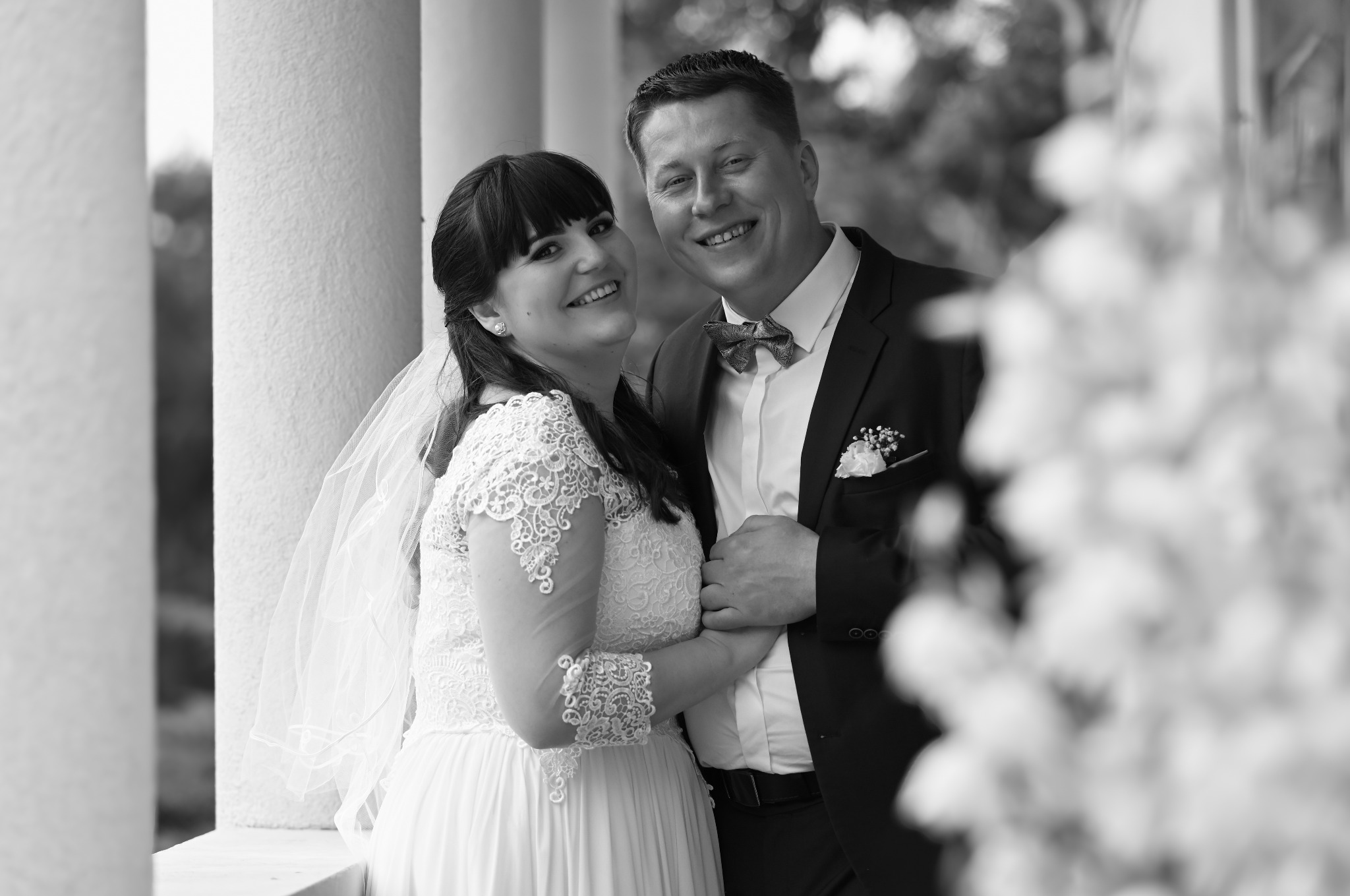 Czarno-białe zdjęcie uśmiechniętej pary młodej, stojącej przy kolumnach. Panna młoda w koronkowej sukni, pan młody w garniturze z muszką. Romantyczna atmosfera.