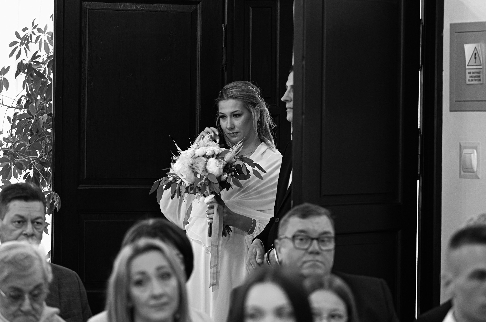 Czarno-białe zdjęcie panny młodej z bukietem, stojącej obok pana młodego na tle ciemnych drzwi, z widocznymi gośćmi weselnymi w pierwszym planie.
