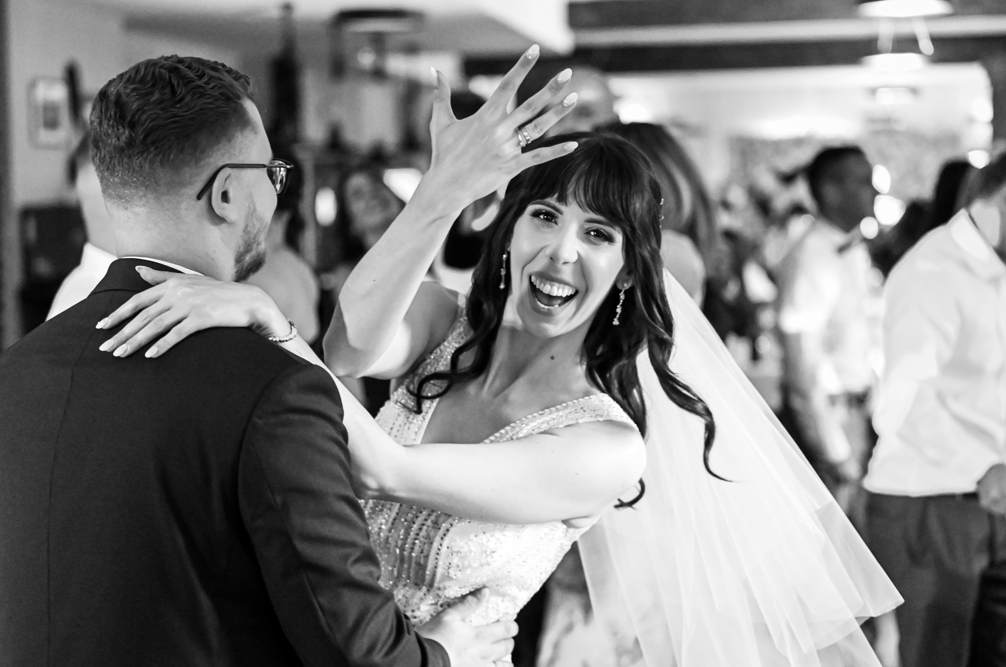 Czarno-białe zdjęcie rozpromienionej panny młodej w objęciach pana młodego, unoszącej dłoń z pierścionkiem podczas weselnego tańca, w tle goście.