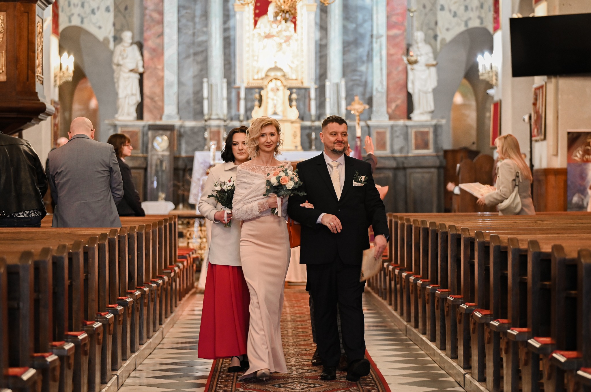 Para młoda w kościele, prowadzona do ołtarza. Panna młoda w jasnej sukni, pan młody w czarnym garniturze. W tle rzędy ławek i ołtarz.