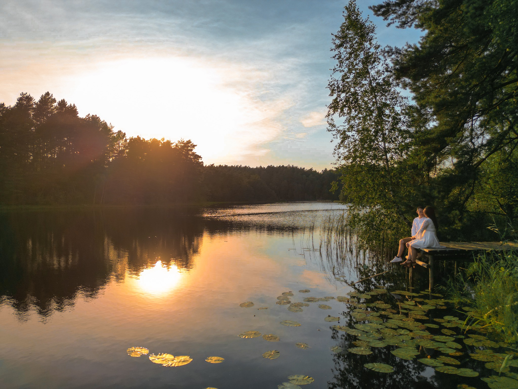 Para siedzi na drewnianym pomoście nad jeziorem o zachodzie słońca. Woda odbija światło, tworząc złote refleksy. Romantyczna sceneria z liliami wodnymi.