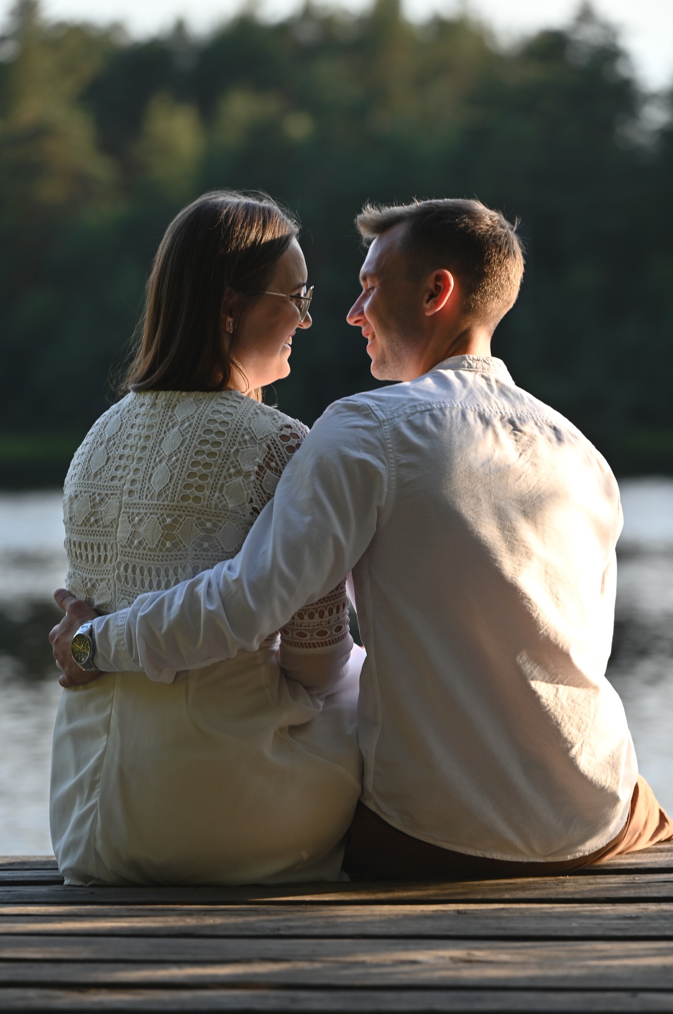 Zakochana para siedzi na drewnianym pomoście nad wodą, patrząc na siebie z uśmiechem. Ona w koronkowej sukience, on w białej koszuli. Romantyczna sesja plenerowa.