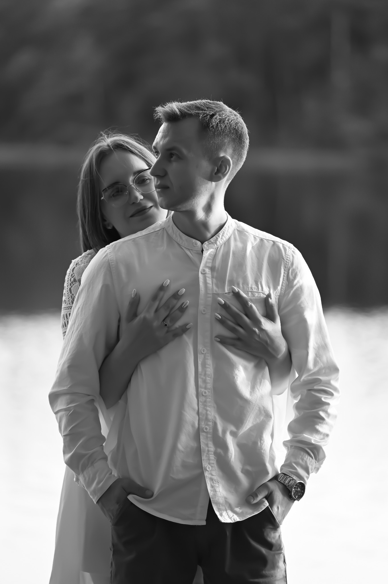 Czarno-białe zdjęcie pary: kobieta w okularach obejmuje mężczyznę w koszuli, oboje patrzą w prawo, na tle rozmytej wody i drzew. Mężczyzna ma zegarek na ręce.