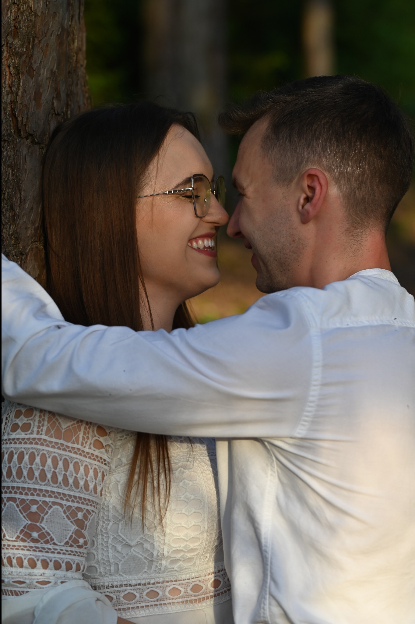 Uśmiechnięta para w objęciach, ona w okularach i koronkowej bluzce, on w białej koszuli, opierają się o drzewo, zbliżenie twarzy, ciepłe światło.
