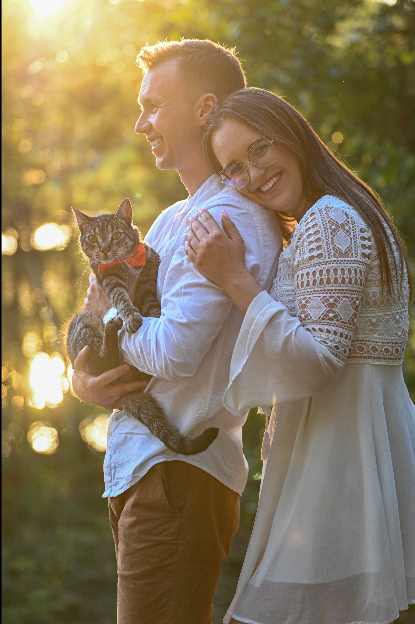 Uśmiechnięta para w plenerze, mężczyzna trzyma kota z czerwoną muchą, kobieta opiera się o niego. Ciepłe, naturalne światło.