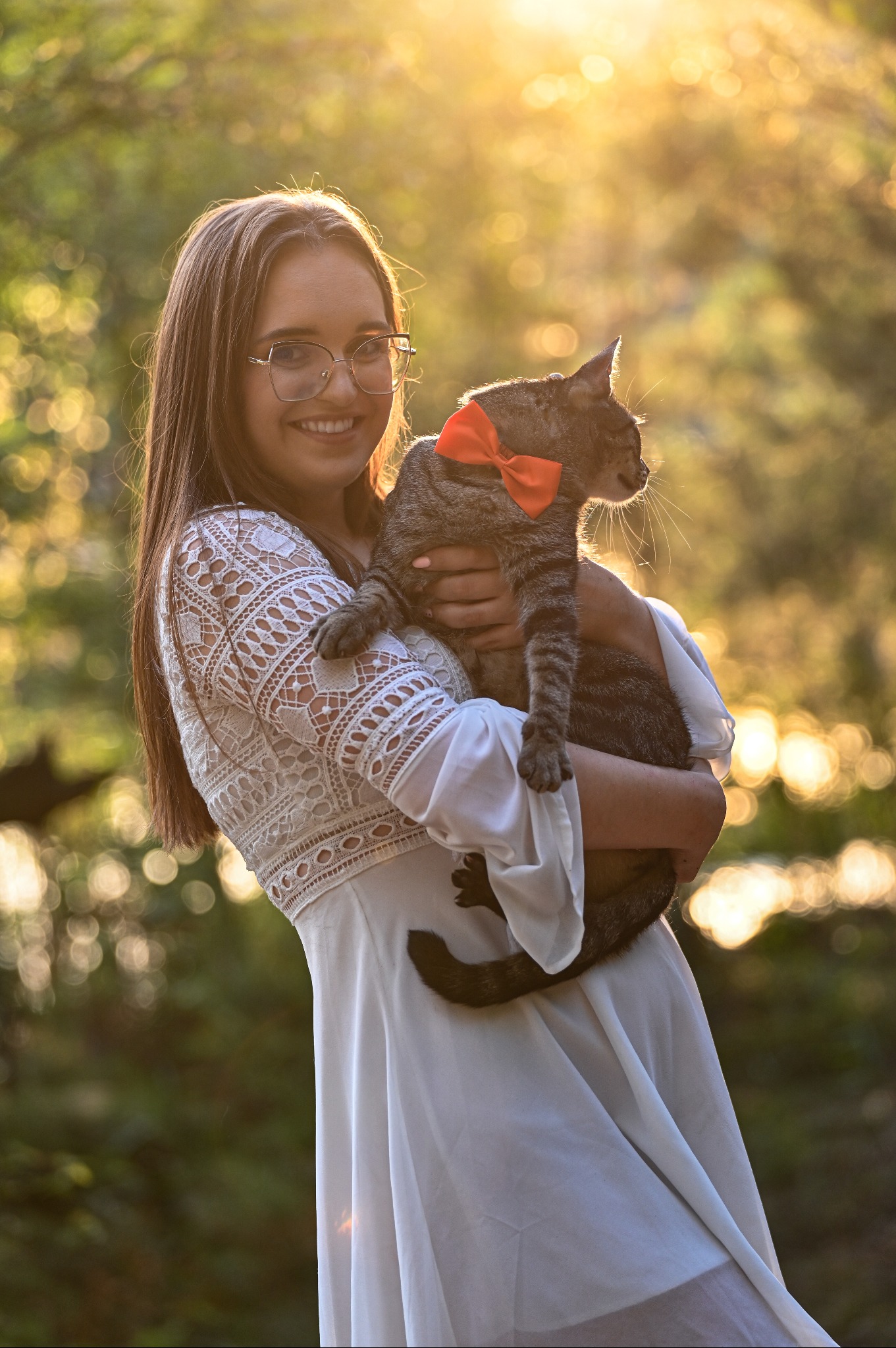 Uśmiechnięta kobieta w okularach trzyma kota z czerwoną kokardą na szyi, w tle rozmyte, słoneczne drzewa. Portret w ciepłym świetle.