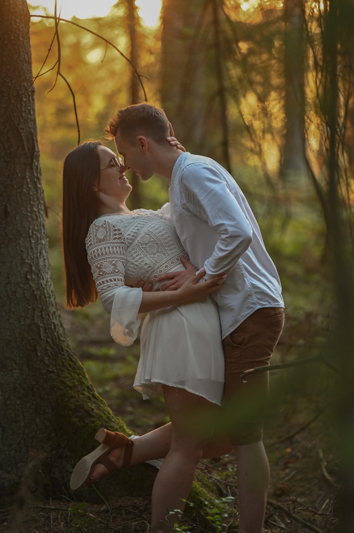 Zakochana para w lesie, ona w białej sukience z koronką, on w białej koszuli i brązowych spodniach, delikatne światło słoneczne, romantyczna sesja w plenerze.