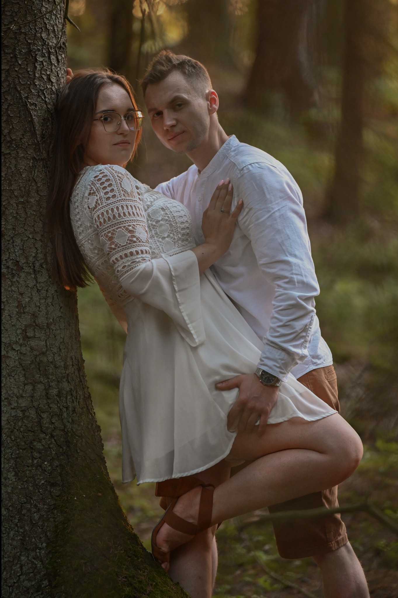 Zakochana para w leśnej scenerii: kobieta w koronkowej sukience opiera się o drzewo, mężczyzna obejmuje ją, patrząc w obiektyw. Romantyczna sesja w naturalnym świetle.