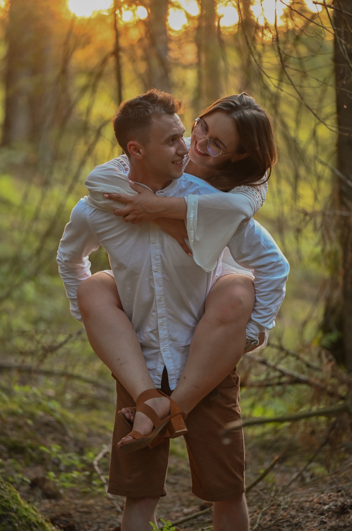 Uśmiechnięta kobieta w okularach na barana u mężczyzny w lesie, ciepłe światło zachodzącego słońca, swobodna atmosfera, naturalne tło.