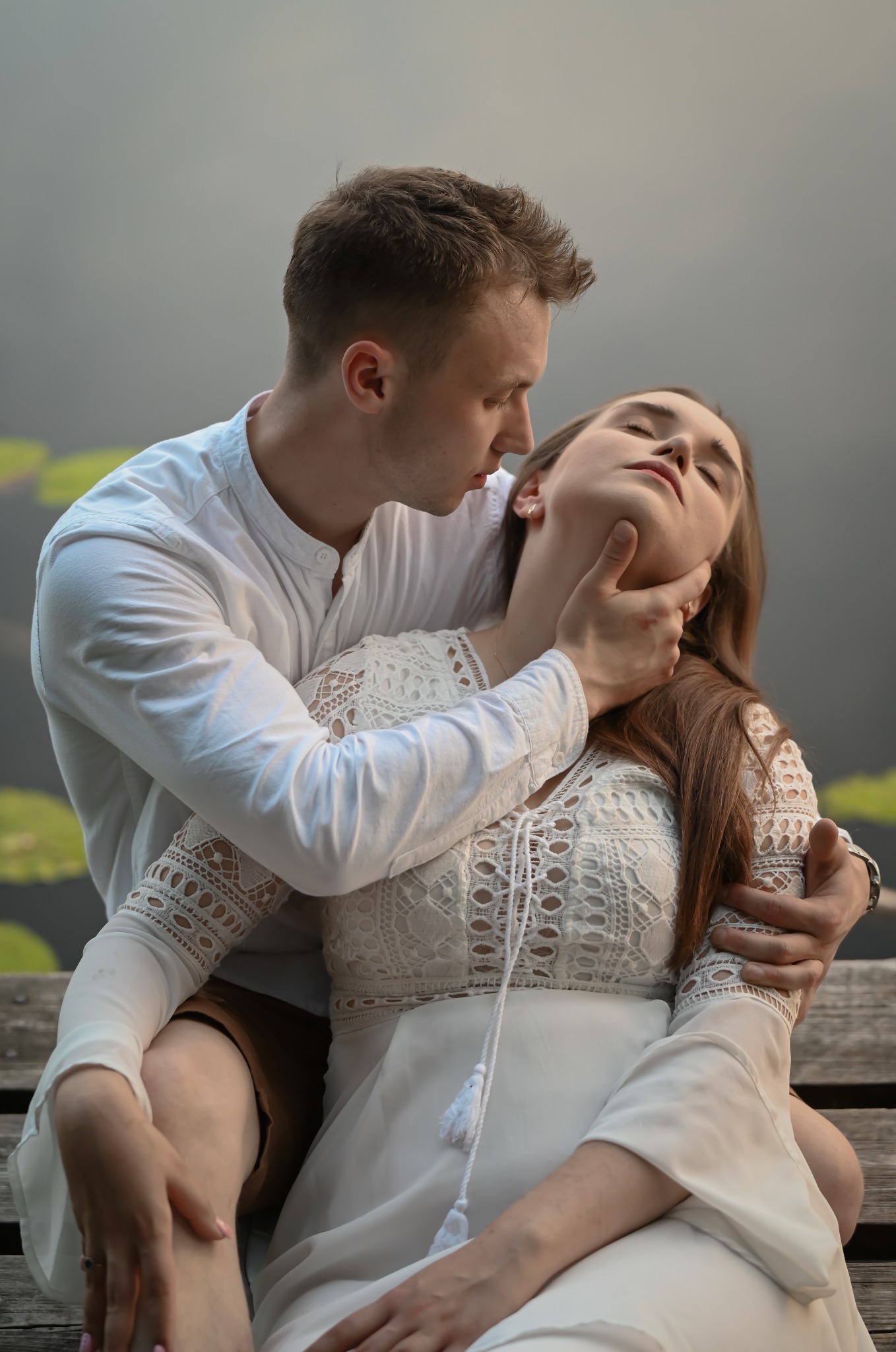 Romantyczna sesja plenerowa: zakochana para w białych strojach siedzi na drewnianym pomoście nad wodą z liliami, delikatne światło podkreśla intymność chwili.