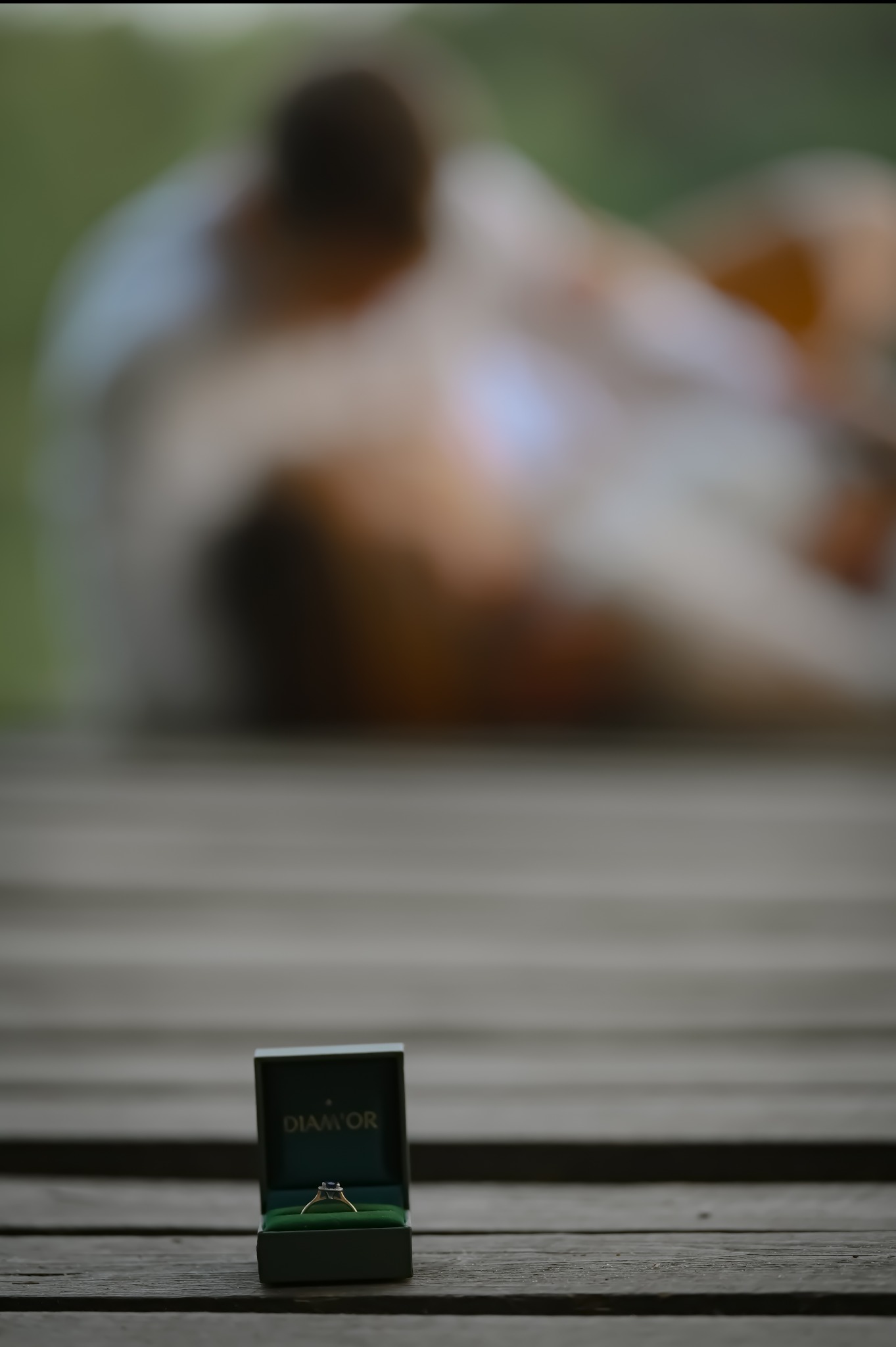 Złoty pierścionek zaręczynowy w otwartym, zielonym pudełku marki Diamor na drewnianym tle. W tle rozmazana para.