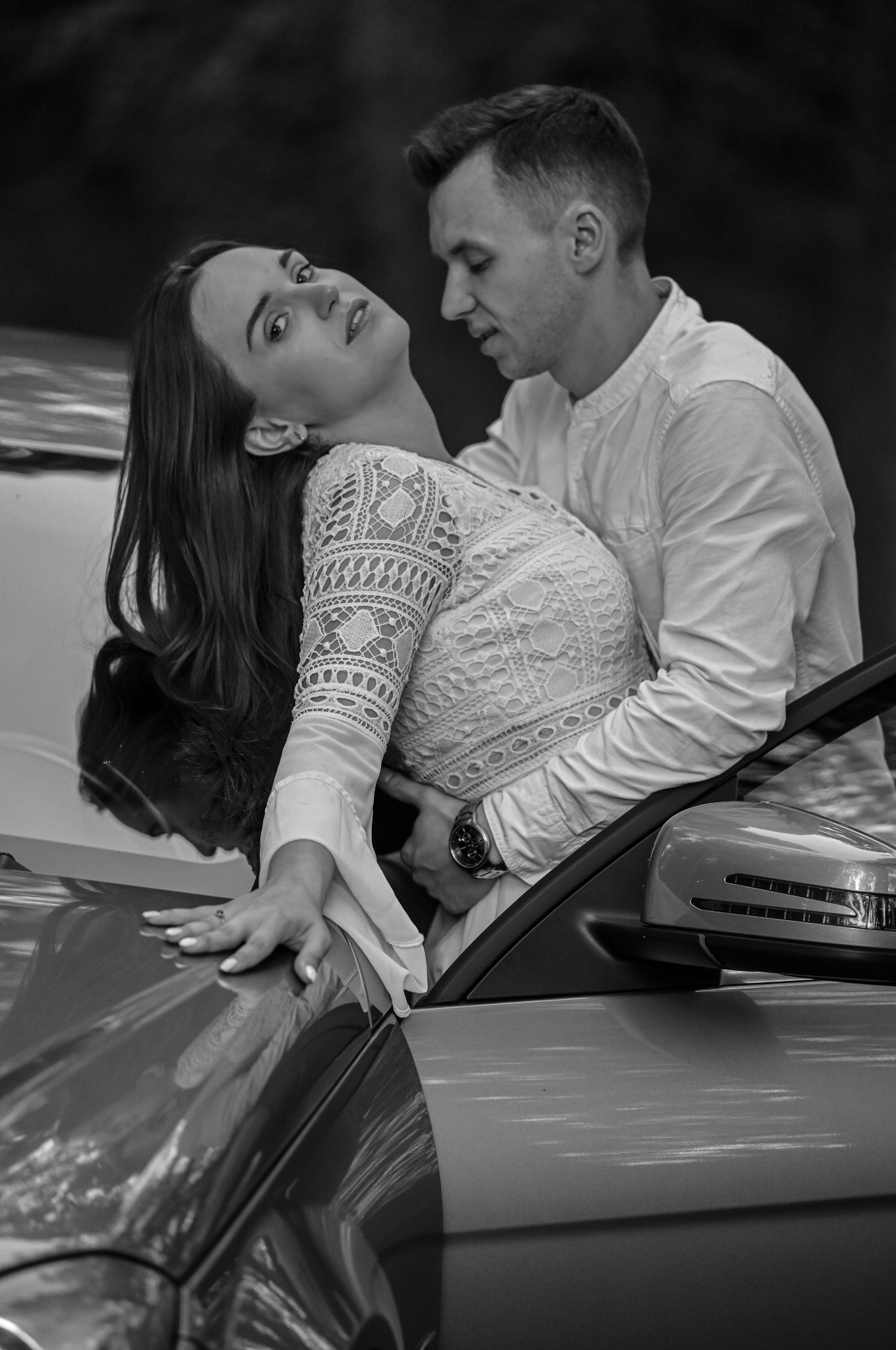 Czarno-białe zdjęcie pary przy samochodzie: kobieta w koronkowej sukience opiera się o maskę, mężczyzna obejmuje ją od tyłu, oboje patrzą w obiektyw.