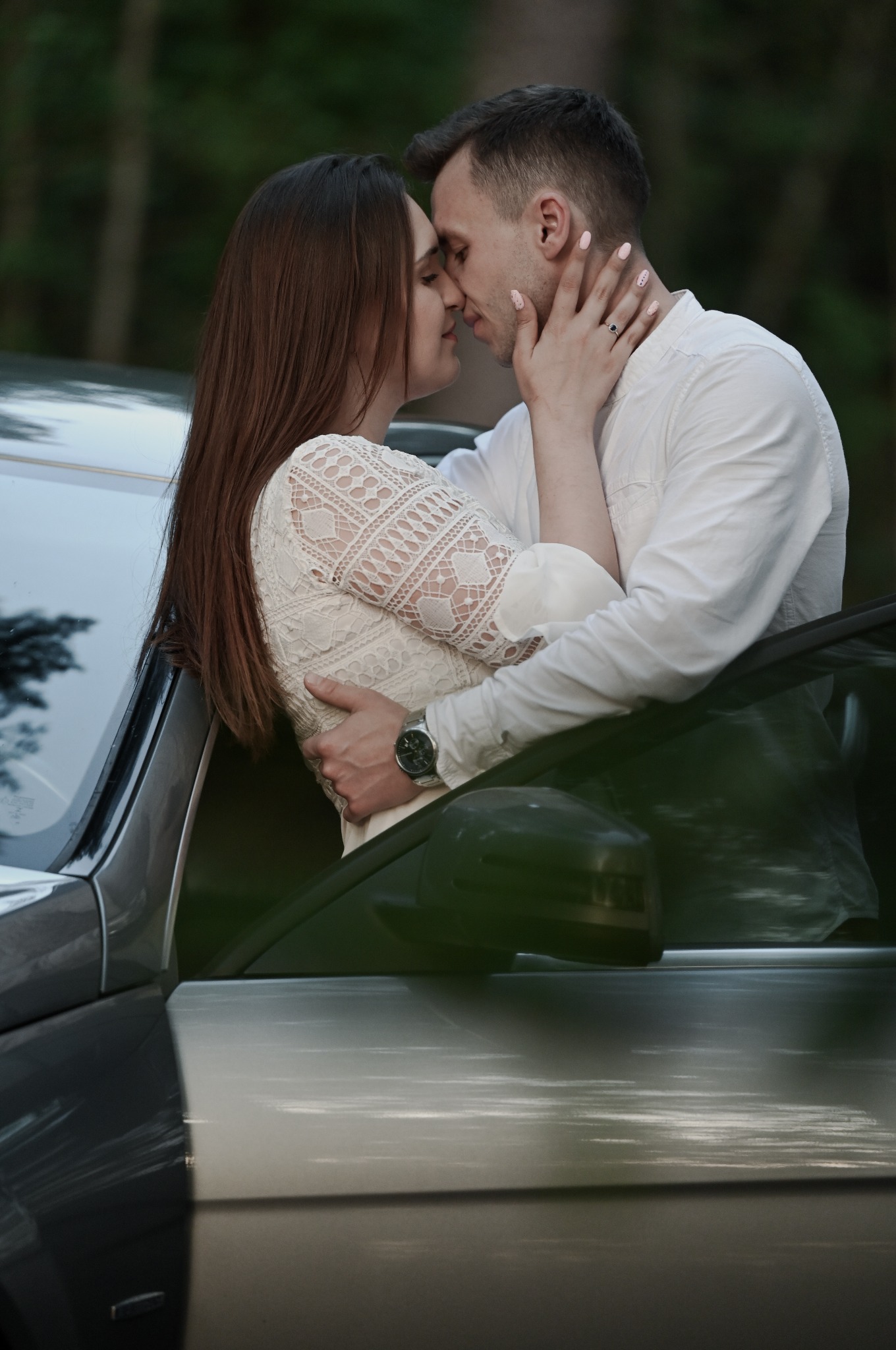 Zakochana para w objęciach, ona w koronkowej sukience, on w białej koszuli, opierają się o samochód. Romantyczna sesja w plenerze, delikatne światło.