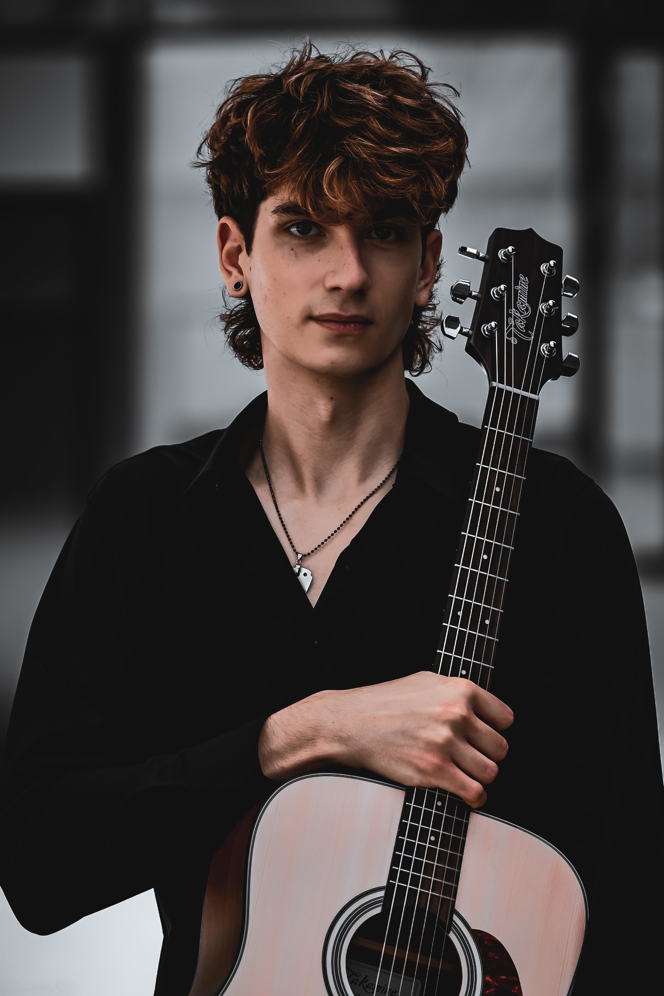 Portret młodego mężczyzny z gitarą akustyczną Takamine. Ubrany w czarną koszulę, z naszyjnikiem. Ujęcie z bliska, z delikatnym rozmyciem tła.