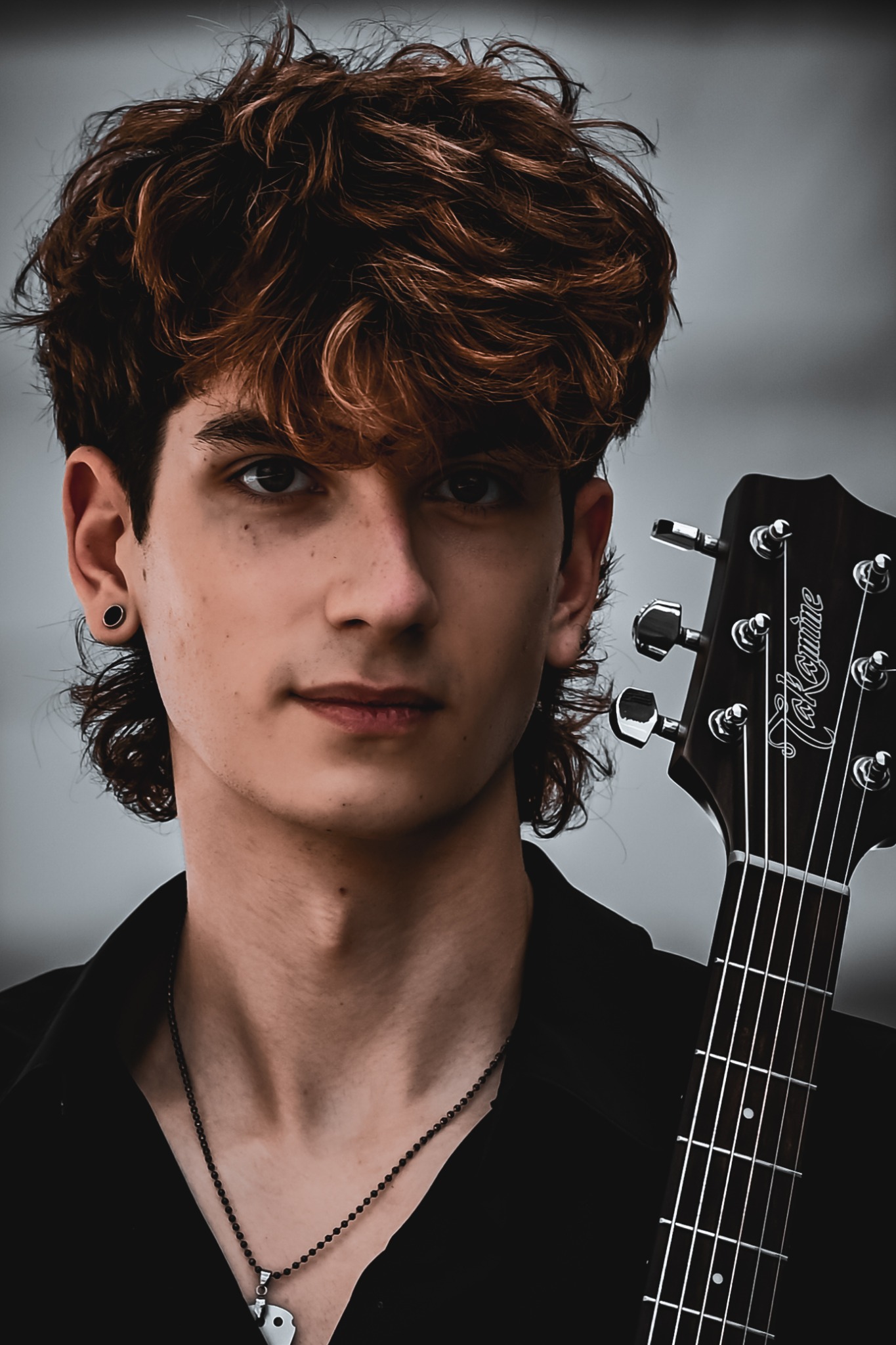 Portret młodego mężczyzny z gitarą Takamine, ciemne włosy z refleksami, czarna koszula i srebrny łańcuszek. Ujęcie z bliska, neutralne tło.