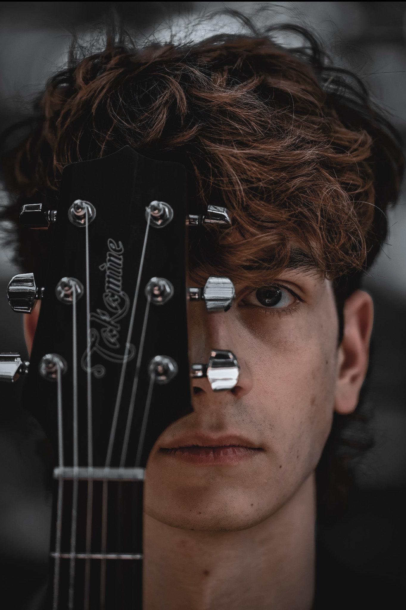Portret młodego mężczyzny z gitarą Takamine, której gryf częściowo zasłania twarz. Widoczne brązowe włosy i jedno oko. Ujęcie w ciemnej tonacji.