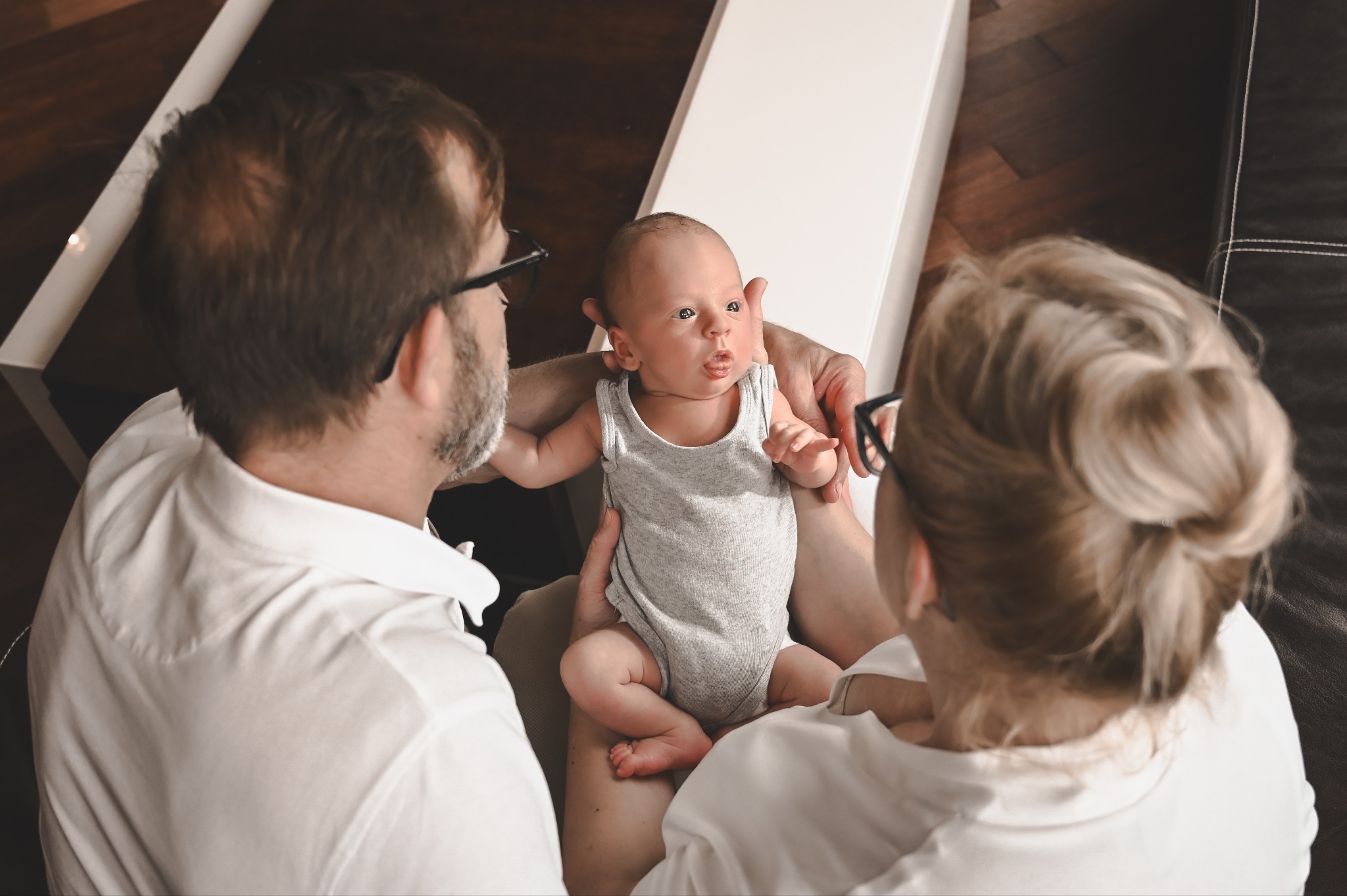 Rodzinne ujęcie z niemowlęciem w centrum, rodzice w białych koszulach patrzą na dziecko. Ciepłe światło, drewniane podłogi w tle. Urocza sesja rodzinna.