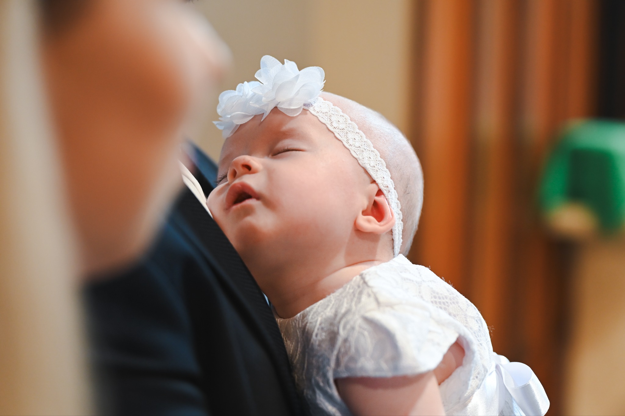 Śpiące niemowlę w białej sukience i opasce z kwiatem, trzymane na rękach osoby w ciemnym garniturze. Ujęcie z bliska, delikatne światło.