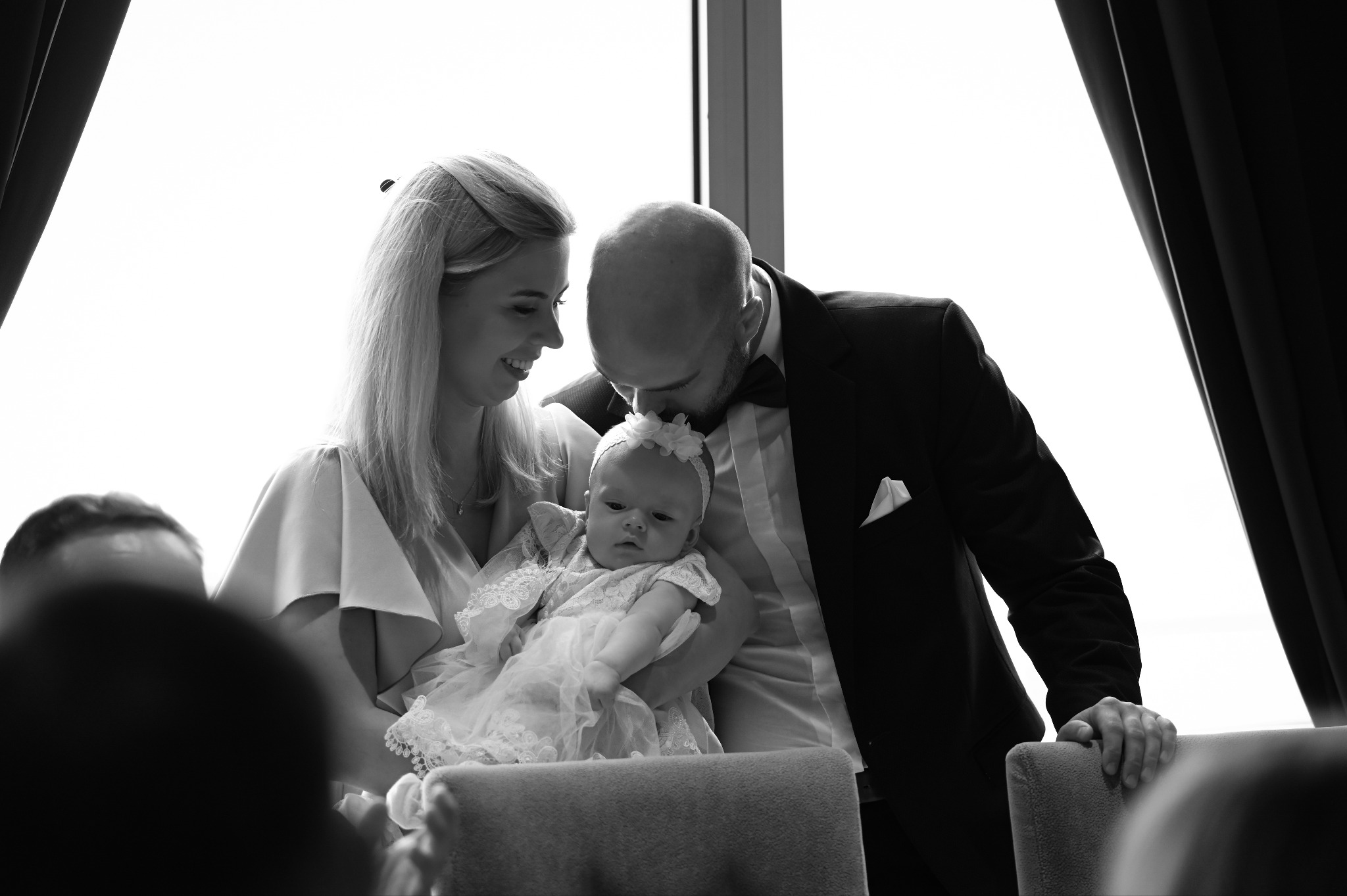 Czarno-białe zdjęcie z Olsztyna: elegancka rodzina z niemowlęciem w sukience z koronką, ojciec całuje dziecko w głowę, jasne tło okna.