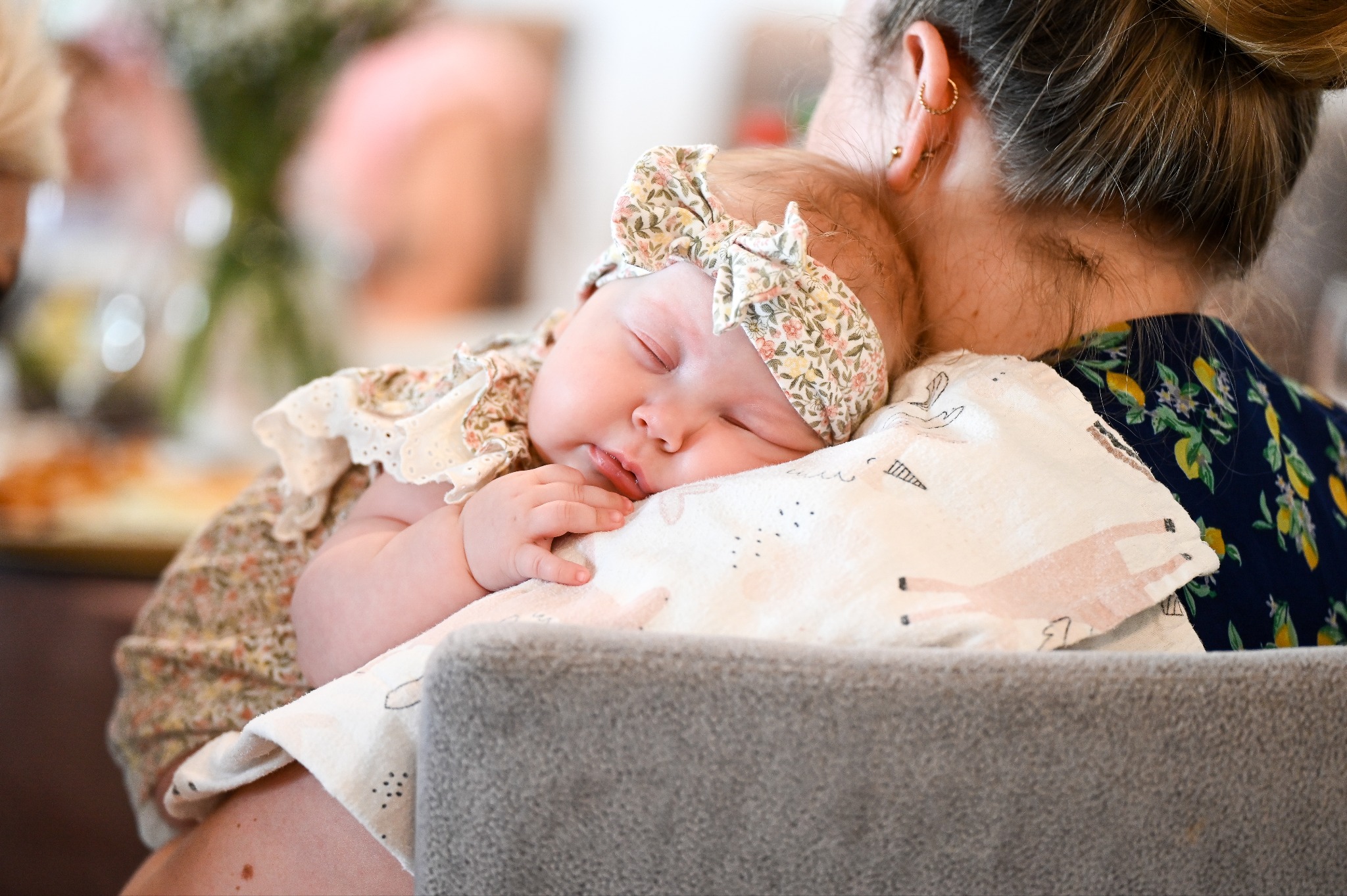 Śpiąca niemowlęca dziewczynka z opaską w kwiaty, wtulona w ramię osoby w kwiecistej bluzce, na tle rozmytych kwiatów.