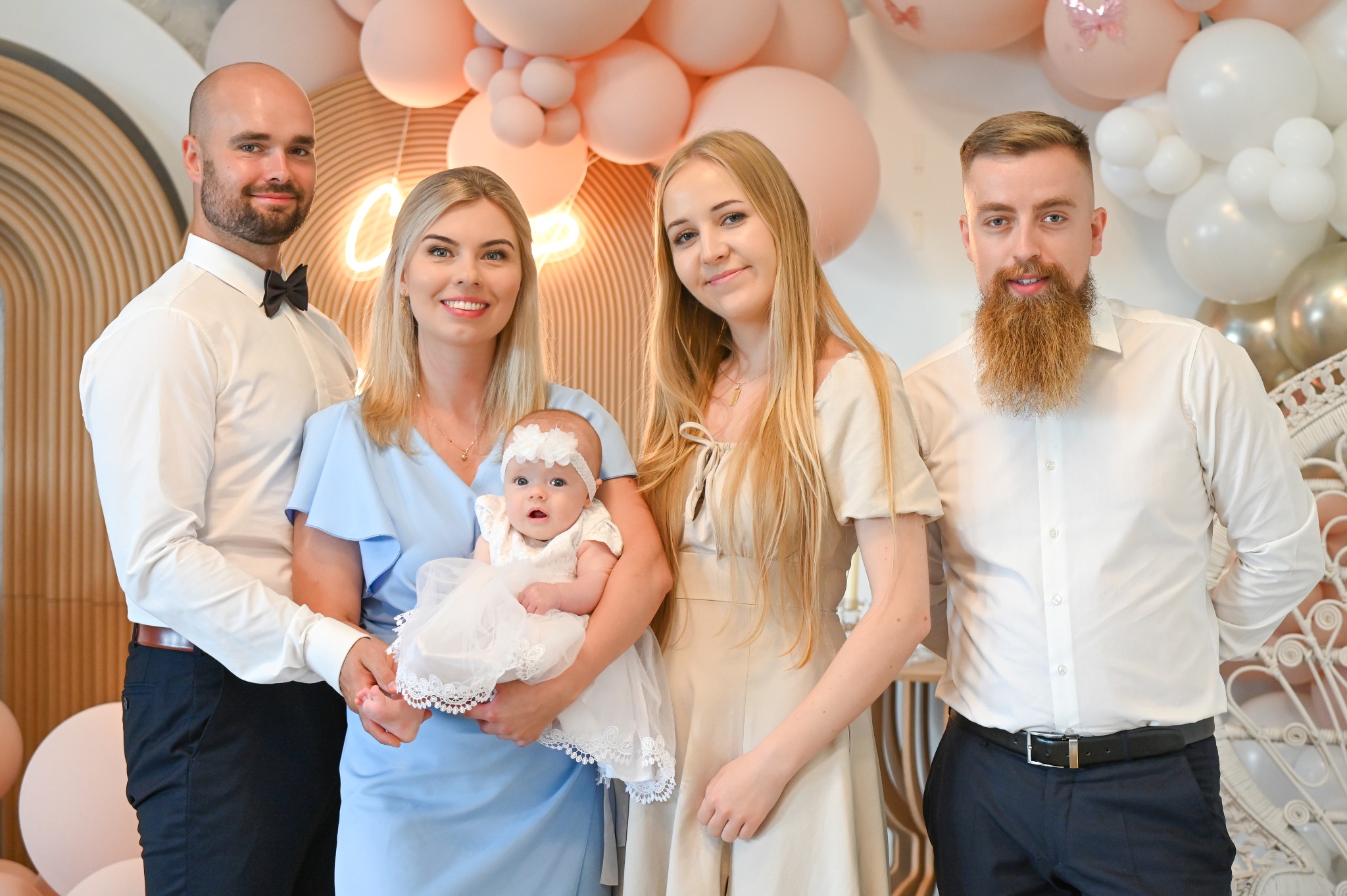 Uśmiechnięta rodzina z niemowlęciem w białej sukience na tle dekoracji z różowych i białych balonów. Mężczyzna w muszce, kobieta w błękitnej sukience. Jasne, naturalne światło.