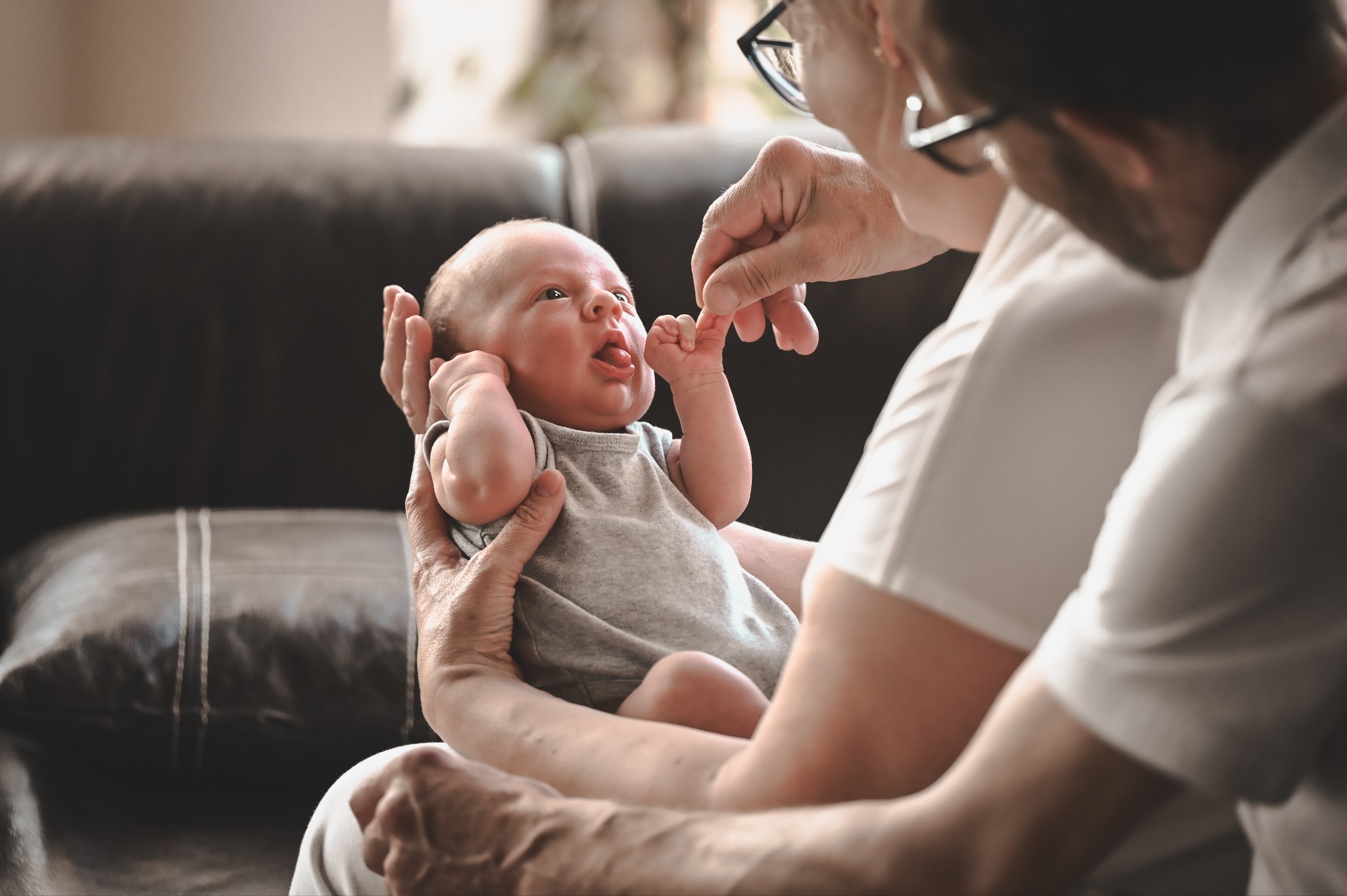 Rodzinne ujęcie: noworodek w szarym ubranku trzymany na rękach przez dorosłych, z delikatnym dotykiem dłoni i miękkim, naturalnym oświetleniem w tle.