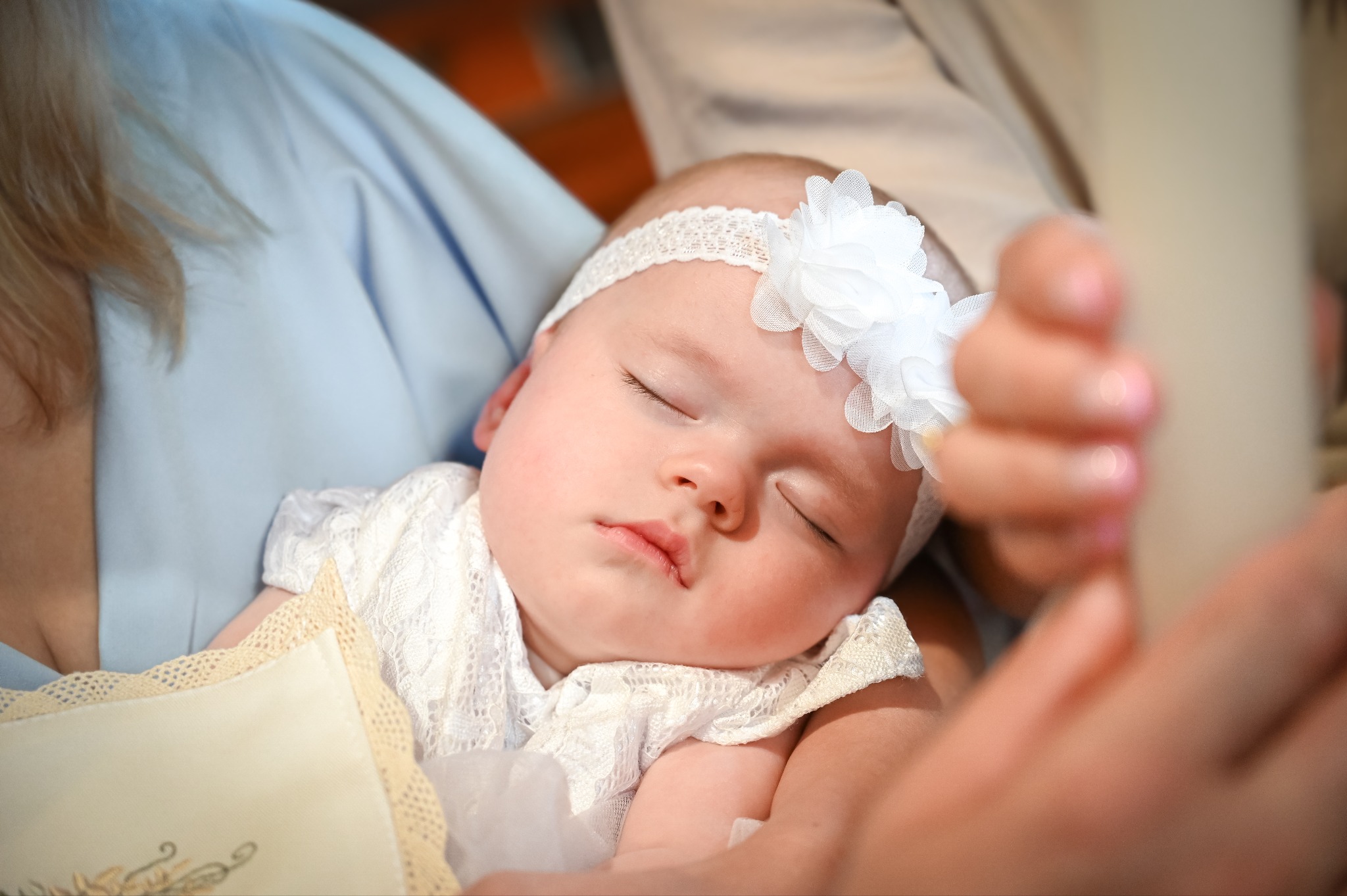 Śpiące niemowlę w białej sukience i opasce z kwiatem, trzymane na rękach. Delikatne światło podkreśla rysy twarzy dziecka. Ujęcie z bliska, skupione na detalu.