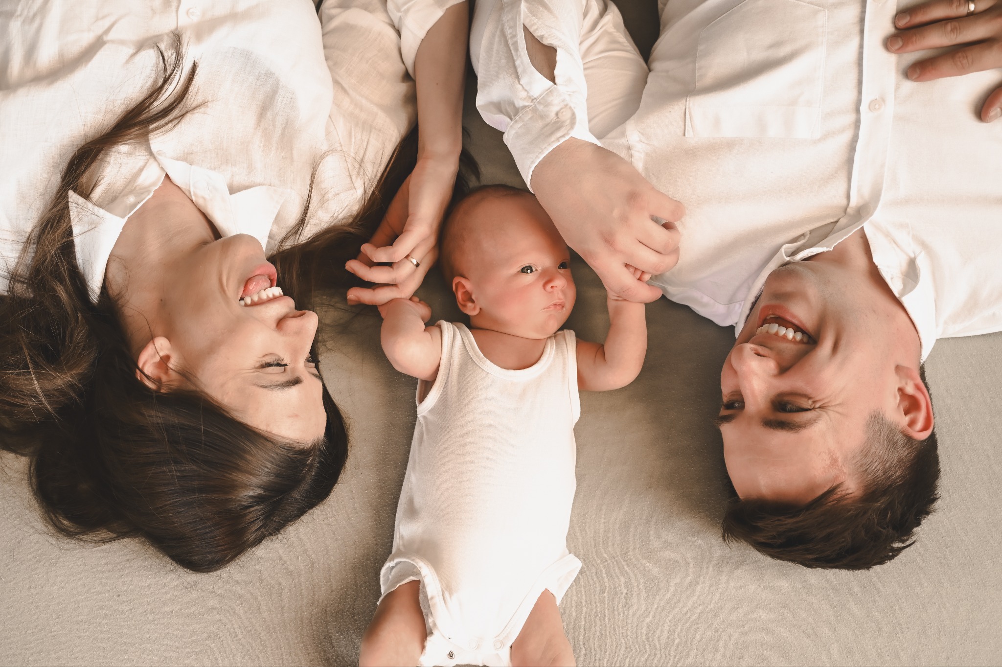 Uśmiechnięta rodzina leżąca na plecach: mama z rozpuszczonymi włosami, tata i niemowlę w białym body trzymające się za ręce. Zdjęcie z góry, ciepłe światło.