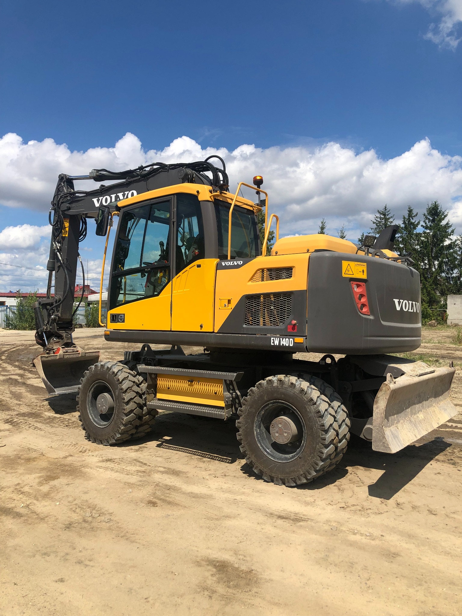 Żółto-szara koparka kołowa Volvo EW140D na placu budowy, z łyżką i lemieszem, na tle błękitnego nieba z chmurami i zieleni drzew.