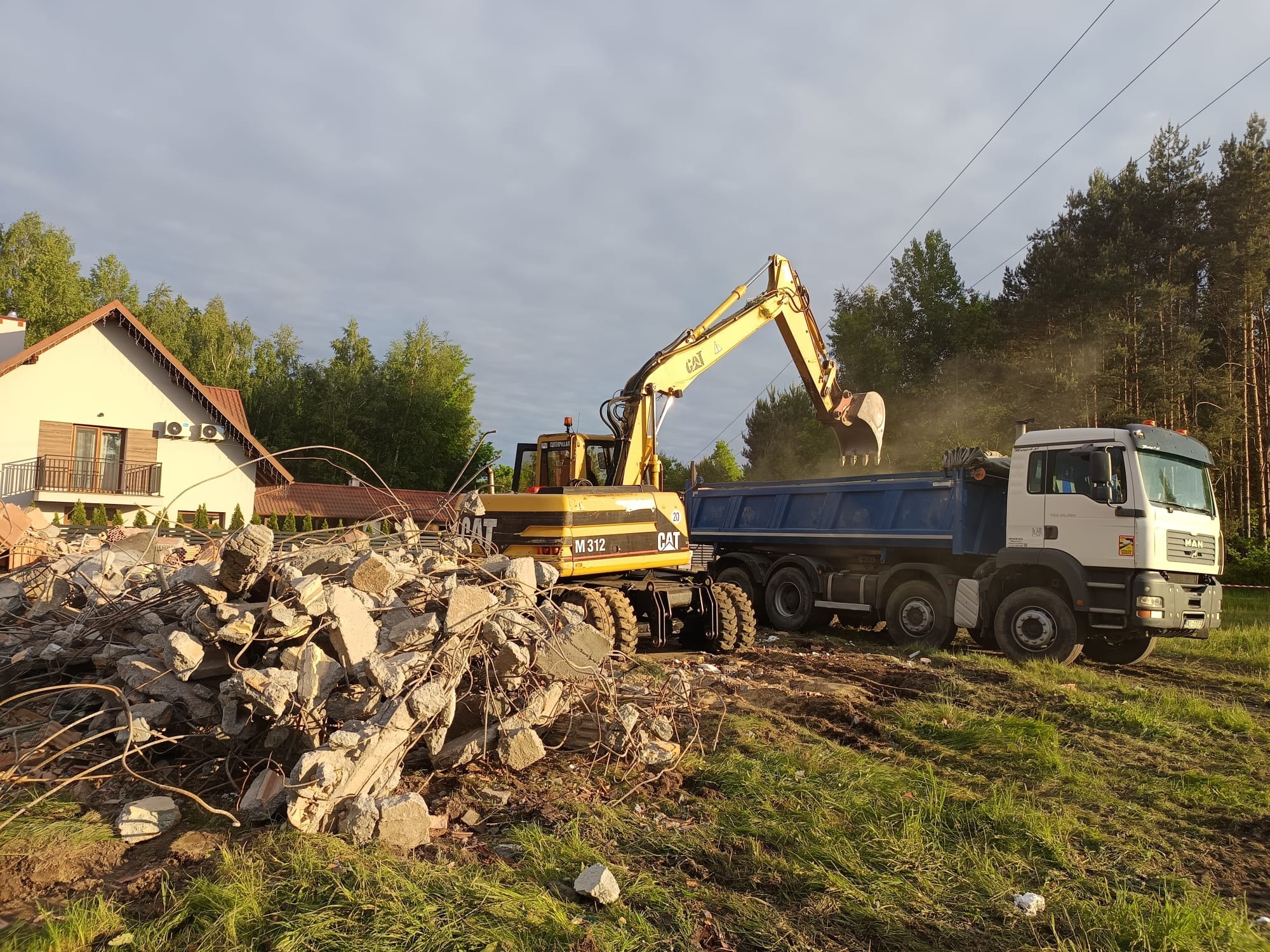 Żółta koparka CAT M312 ładuje gruz z rozbiórki do niebieskiej skrzyni ciężarówki MAN. W tle dom i drzewa.