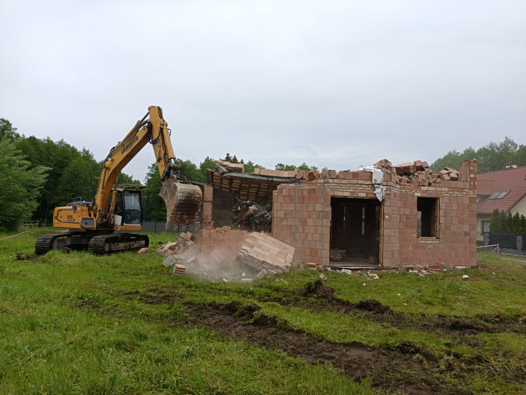 Żółta koparka Liebherr 906 burzy ceglany budynek. Widoczne gruz i pył. W tle zieleń drzew i dom z czerwoną dachówką. Pochmurne niebo.