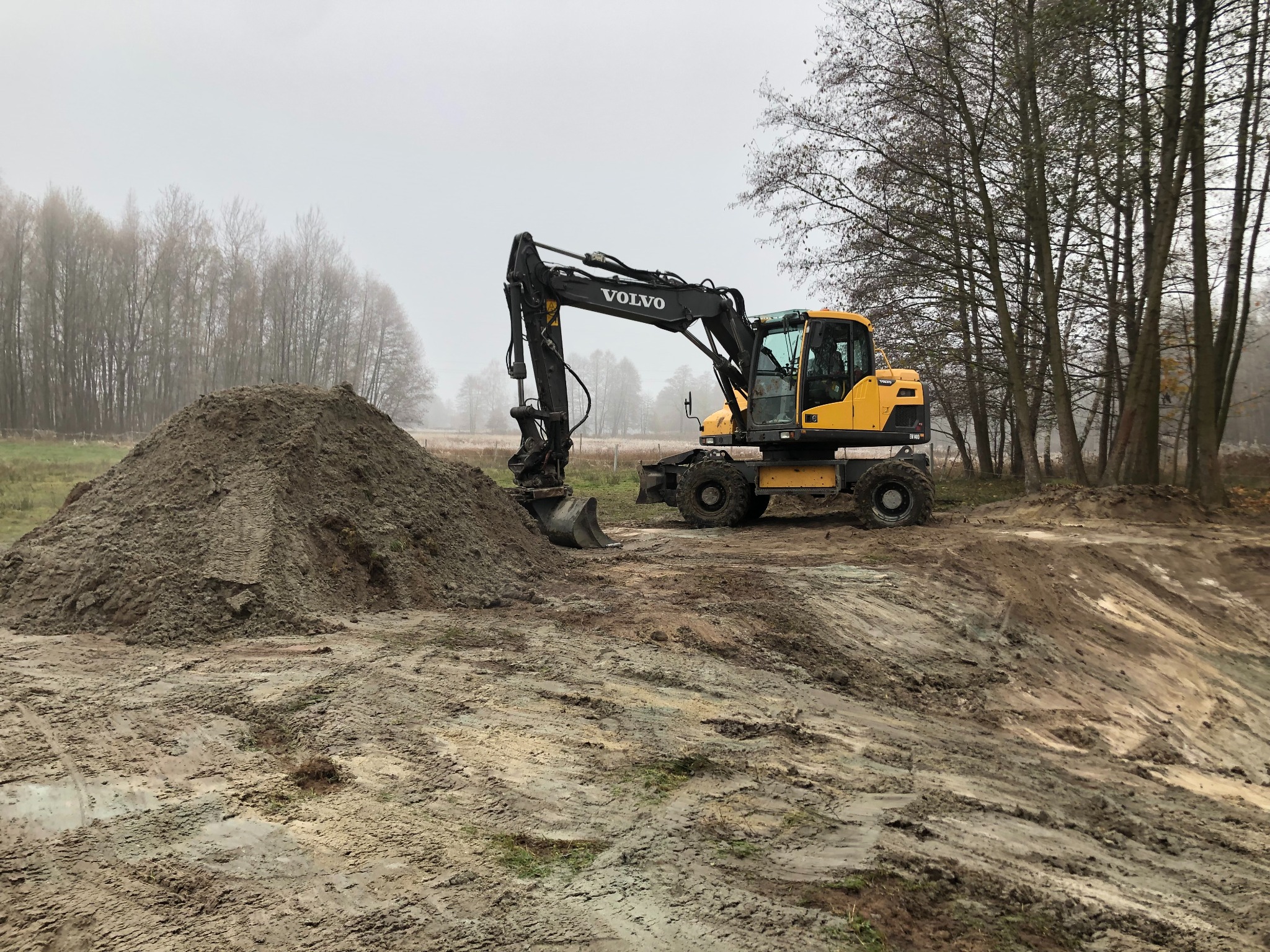 Żółta koparka Volvo na błotnistym terenie z pryzmą ziemi. W tle drzewa i mglista pogoda. Widoczne ślady opon i prace ziemne.