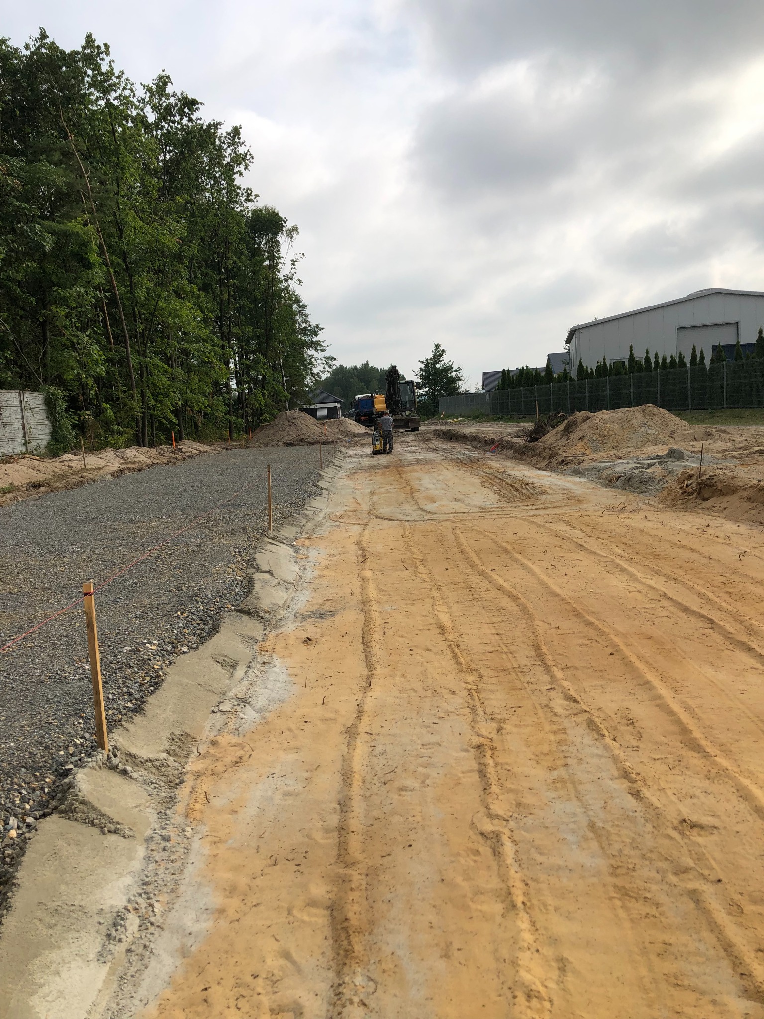 Utwardzanie drogi: wyrównany teren z widocznymi śladami opon, częściowo wysypany żwir, w tle maszyny budowlane i drzewa. Widok na plac budowy w pochmurny dzień.