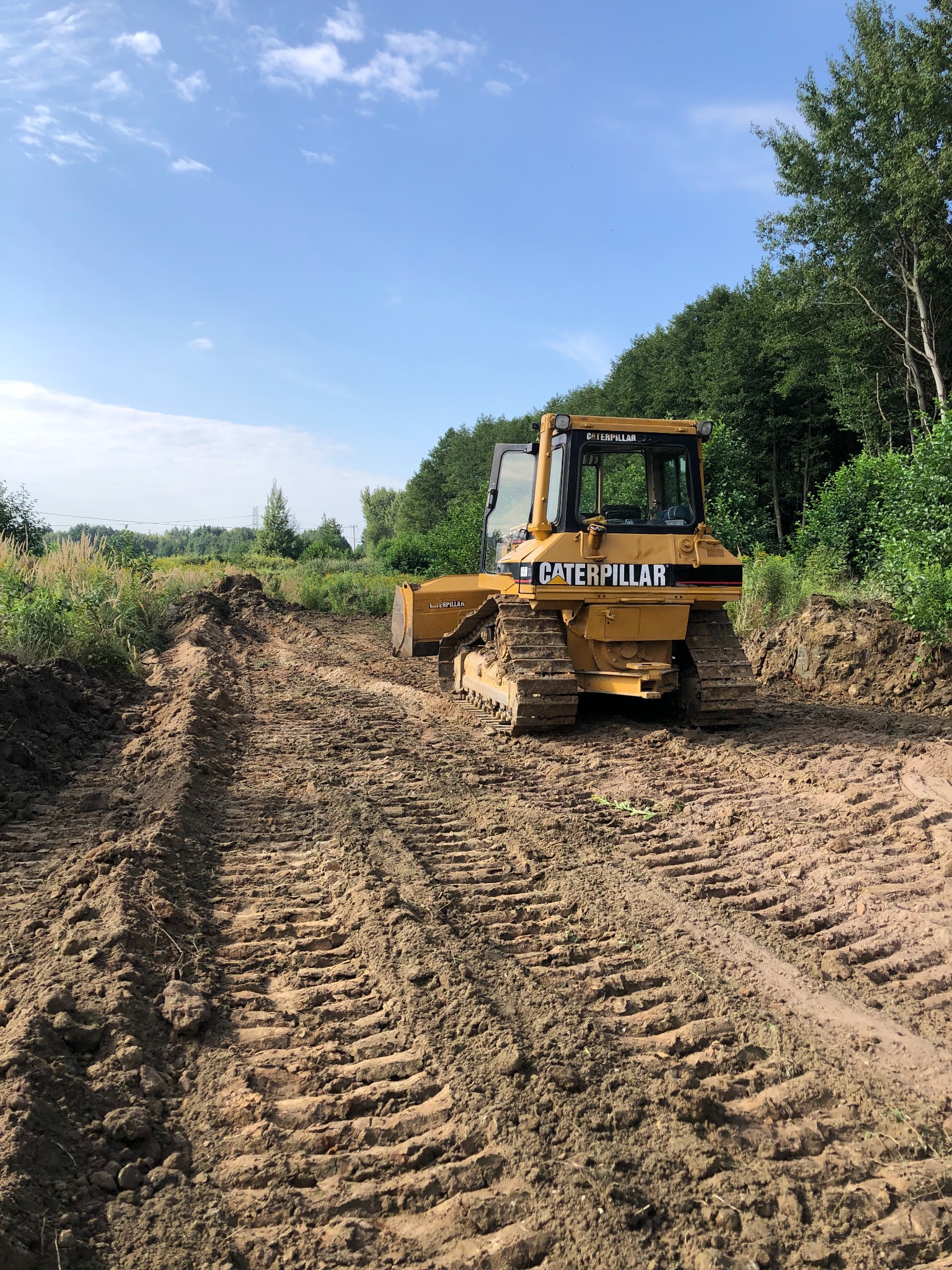 Żółty spychacz Caterpillar na nierównym, błotnistym terenie z wyraźnymi śladami gąsienic, otoczony zielenią i drzewami pod błękitnym niebem.