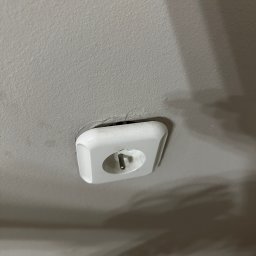 Biuro Inwestycji i Nadzoru Budowlanego - Białe gniazdko elektryczne typu E wystające z jasnego sufitu, widoczne z bliska pod kątem. Widoczne niedoskonałości sufitu wokół gniazdka.