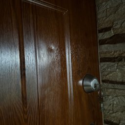 Biuro Inwestycji i Nadzoru Budowlanego - Lakierowane, drewniane drzwi wejściowe z zamkiem i kluczem, widoczne z bliska, z fragmentem kamiennej ściany w tle. Detal wykończenia wnętrza.