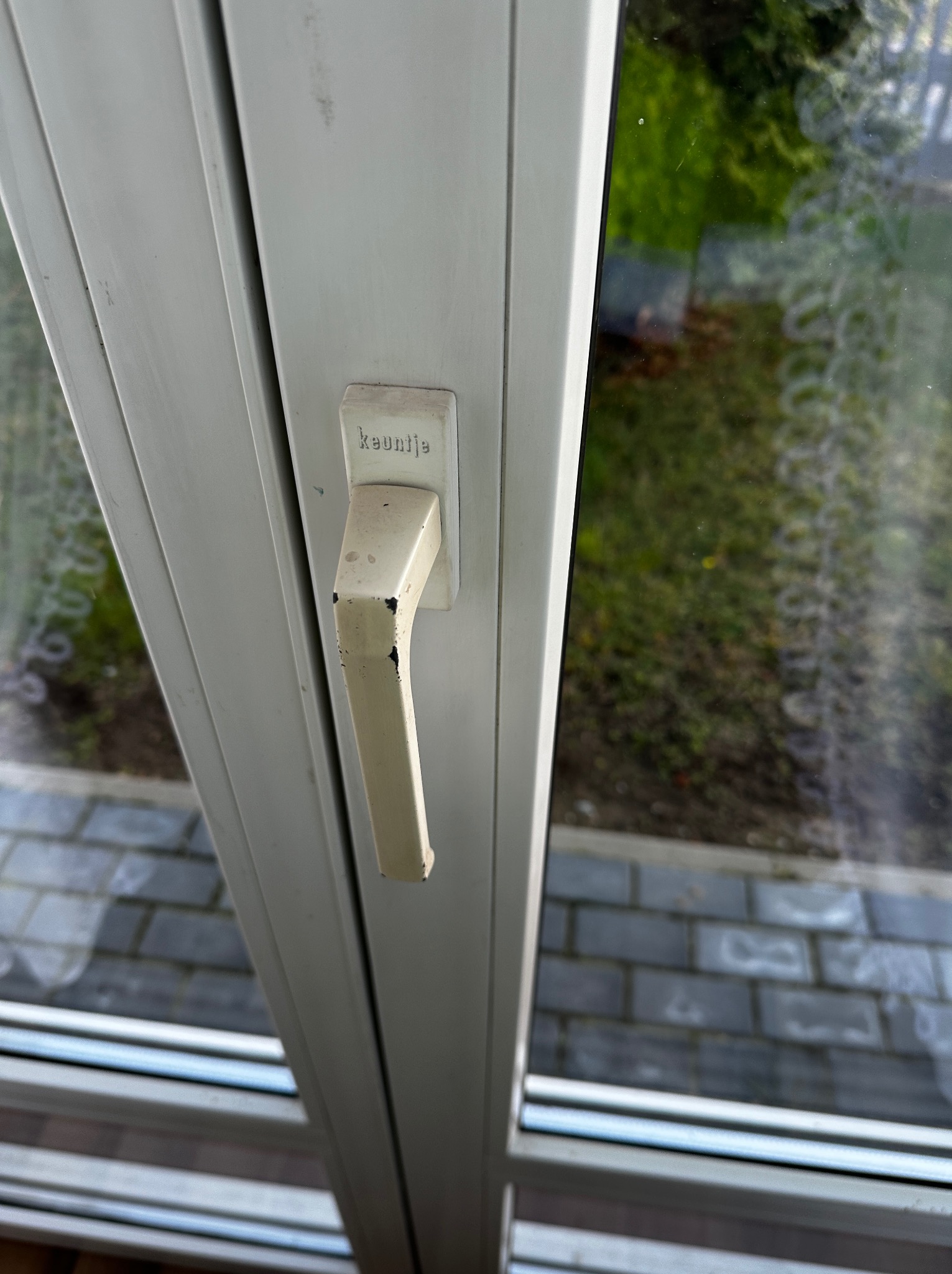 Białe okno PCV z widoczną klamką marki 'keuntje', zarysowaniami i zabrudzeniami. Widok na zewnątrz, na kostkę brukową i zieleń. Ujęcie z bliska, ukazujące detale.