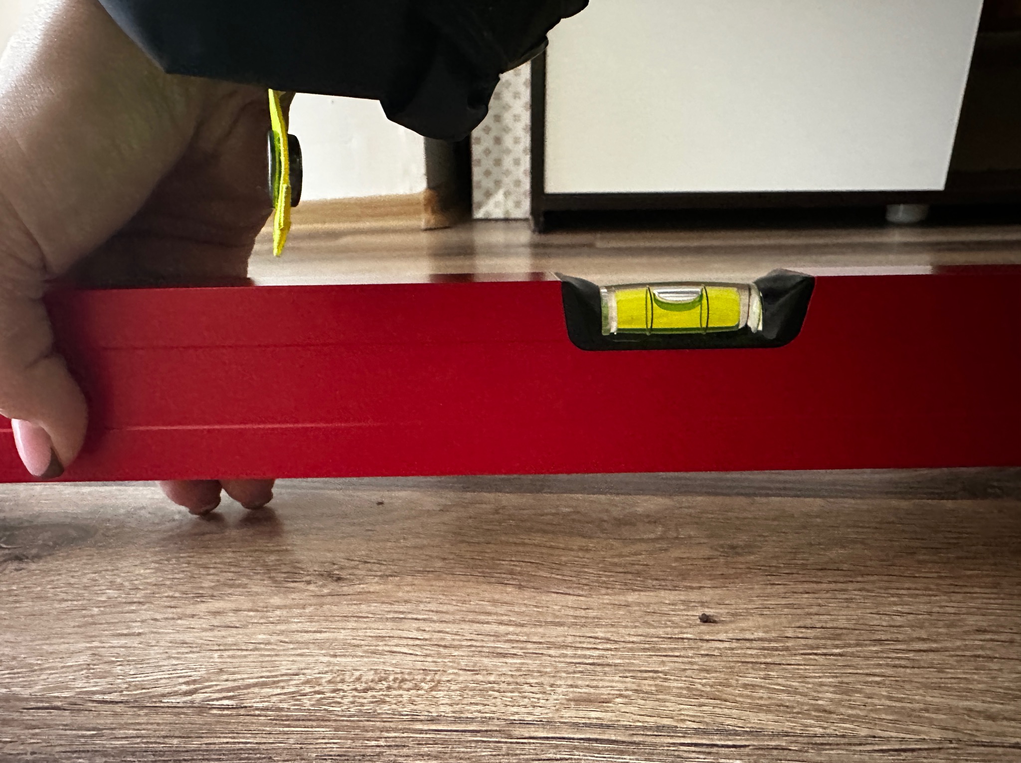Czerwona poziomica w dłoni, sprawdzająca wypoziomowanie drewnianej podłogi. W tle fragment mebla.
