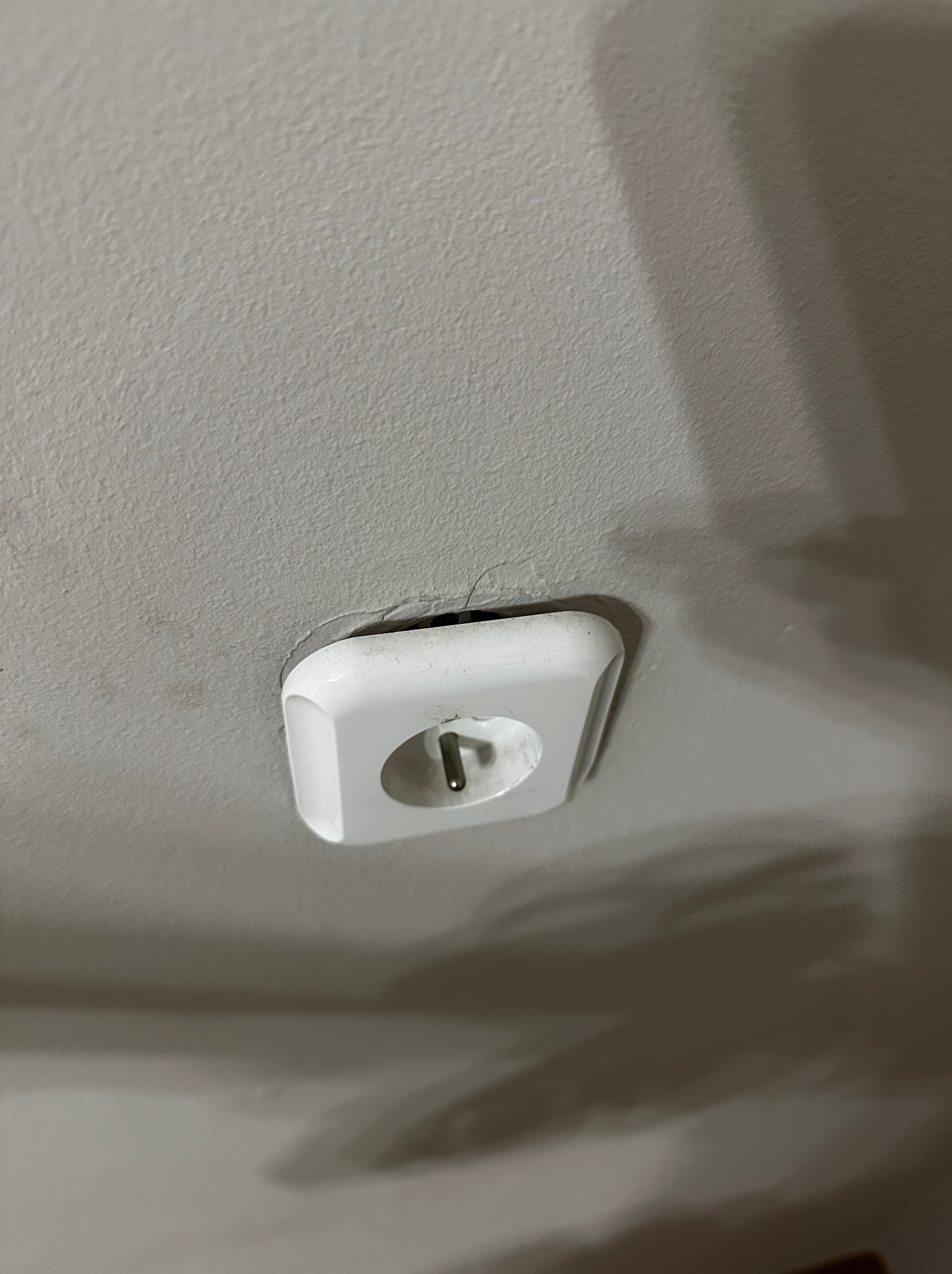 Białe gniazdko elektryczne typu E wystające z jasnego sufitu, widoczne z bliska pod kątem. Widoczne niedoskonałości sufitu wokół gniazdka.